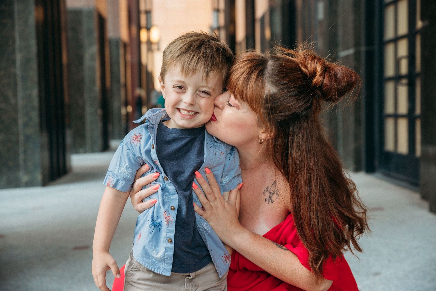 A mother with red hair kisses her smiling young boy on the cheek outdoors in an urban setting.
