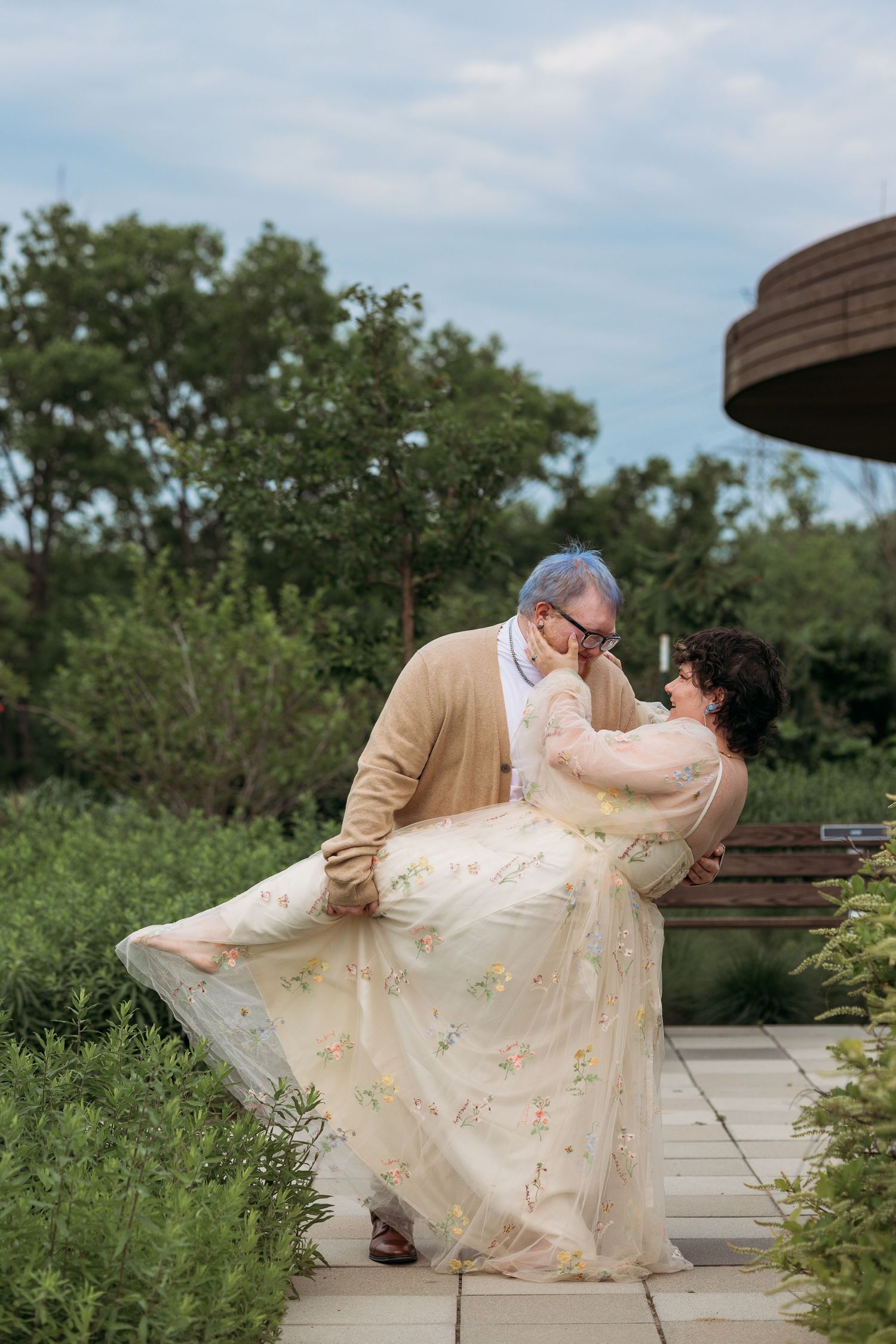 Couple sharing a romantic moment outdoors, man lifting woman in floral dress on garden path with lush greenery backdrop.