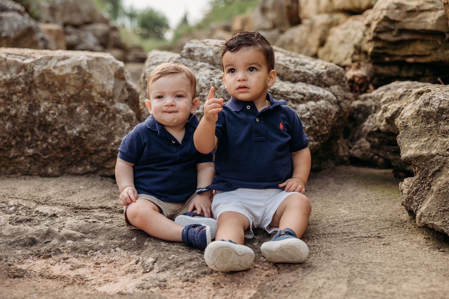 Two baby boys in matching navy polo shirts and white shorts sitting on rocks outdoors.