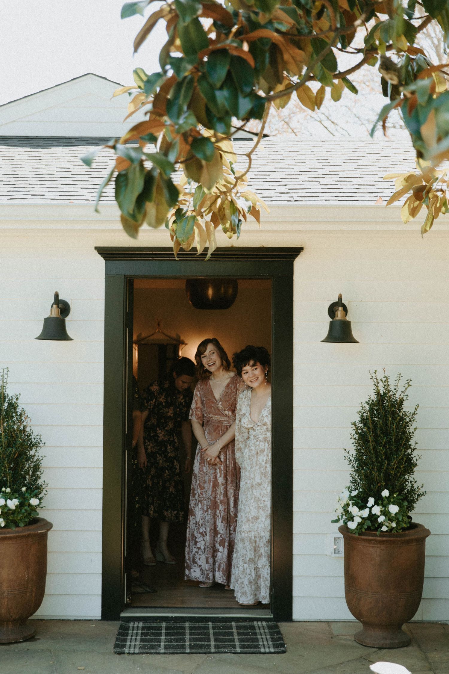 Two people sharing laughter in doorway of white house with potted plants.