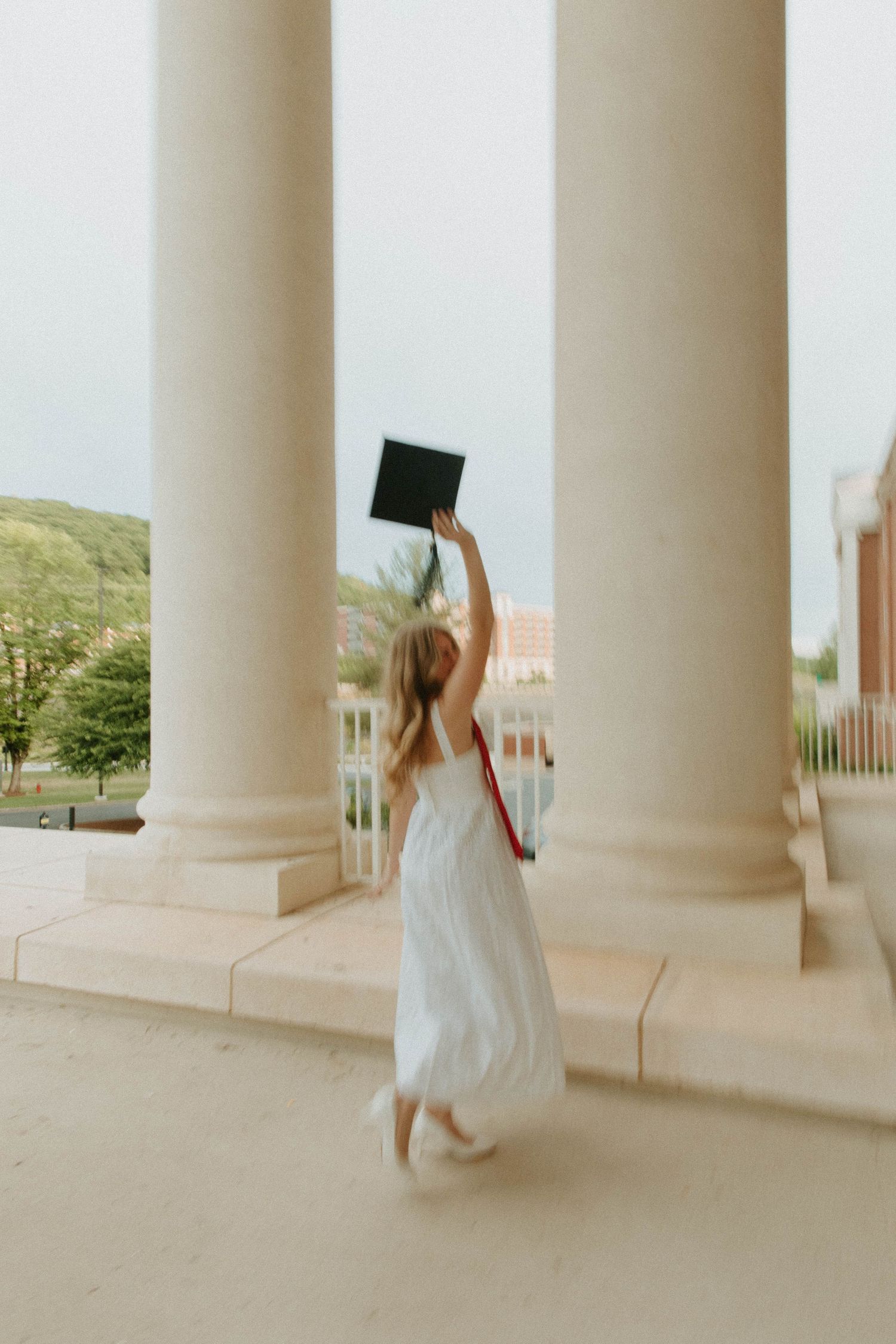 A graduate in a white dress twirls joyfully with their cap held high between classic marble columns.