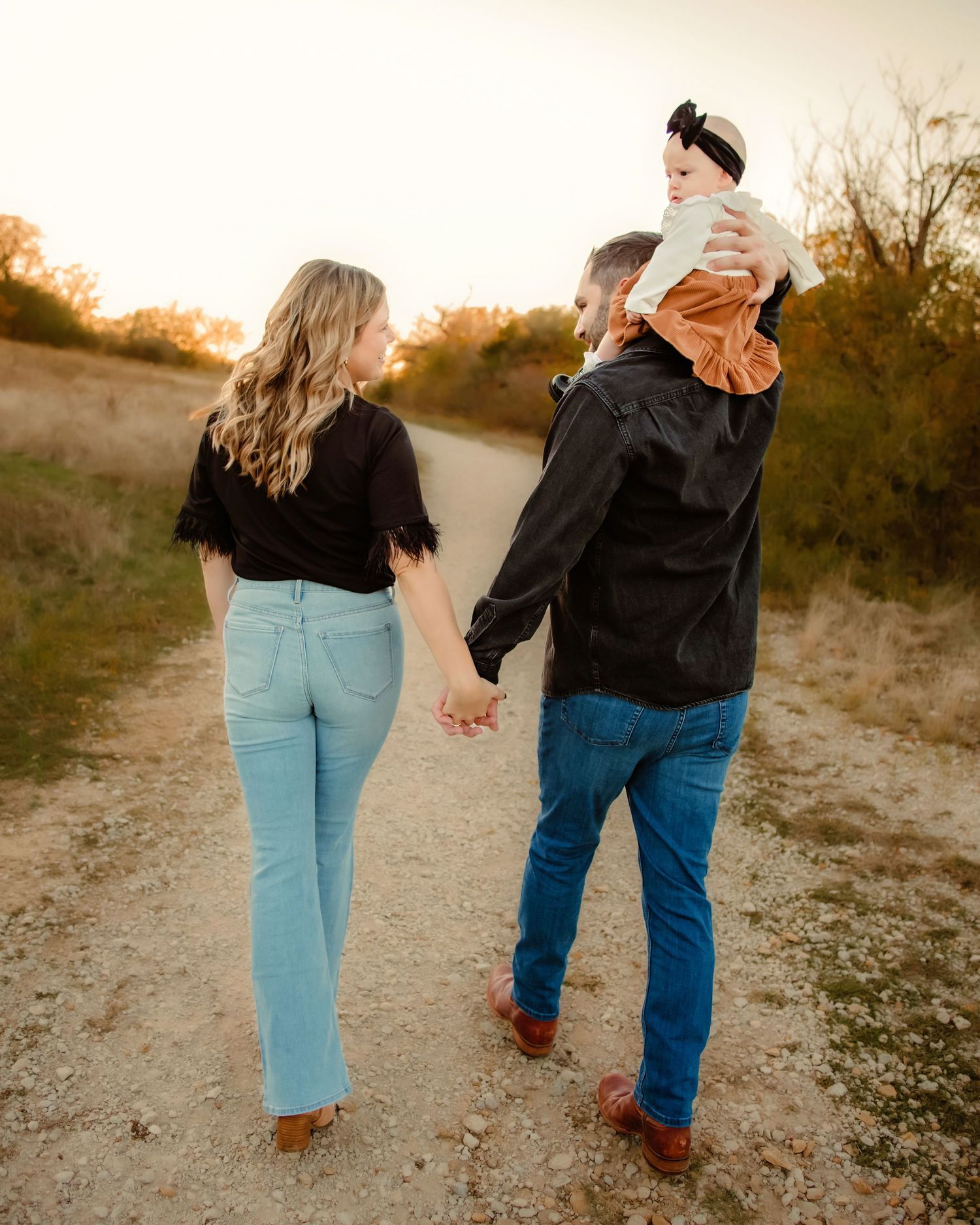 A couple walking hand in hand on a dirt path with their baby riding dad's shoulders during golden hour sunset in Fort Worth, TX.
