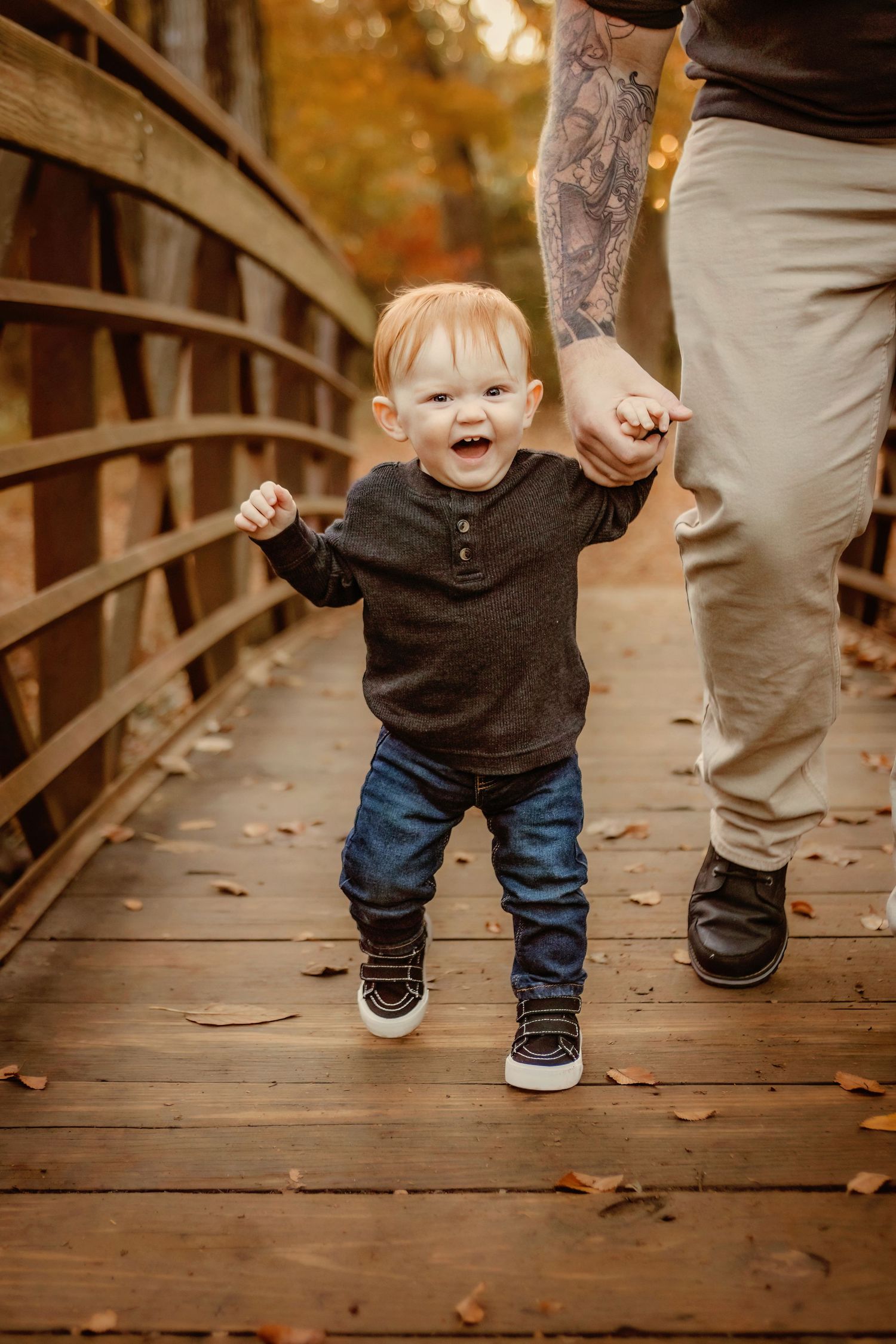 Joyful toddler walking on wooden bridge while holding dad's tattooed hand during family portraits in Colleyville, TX.