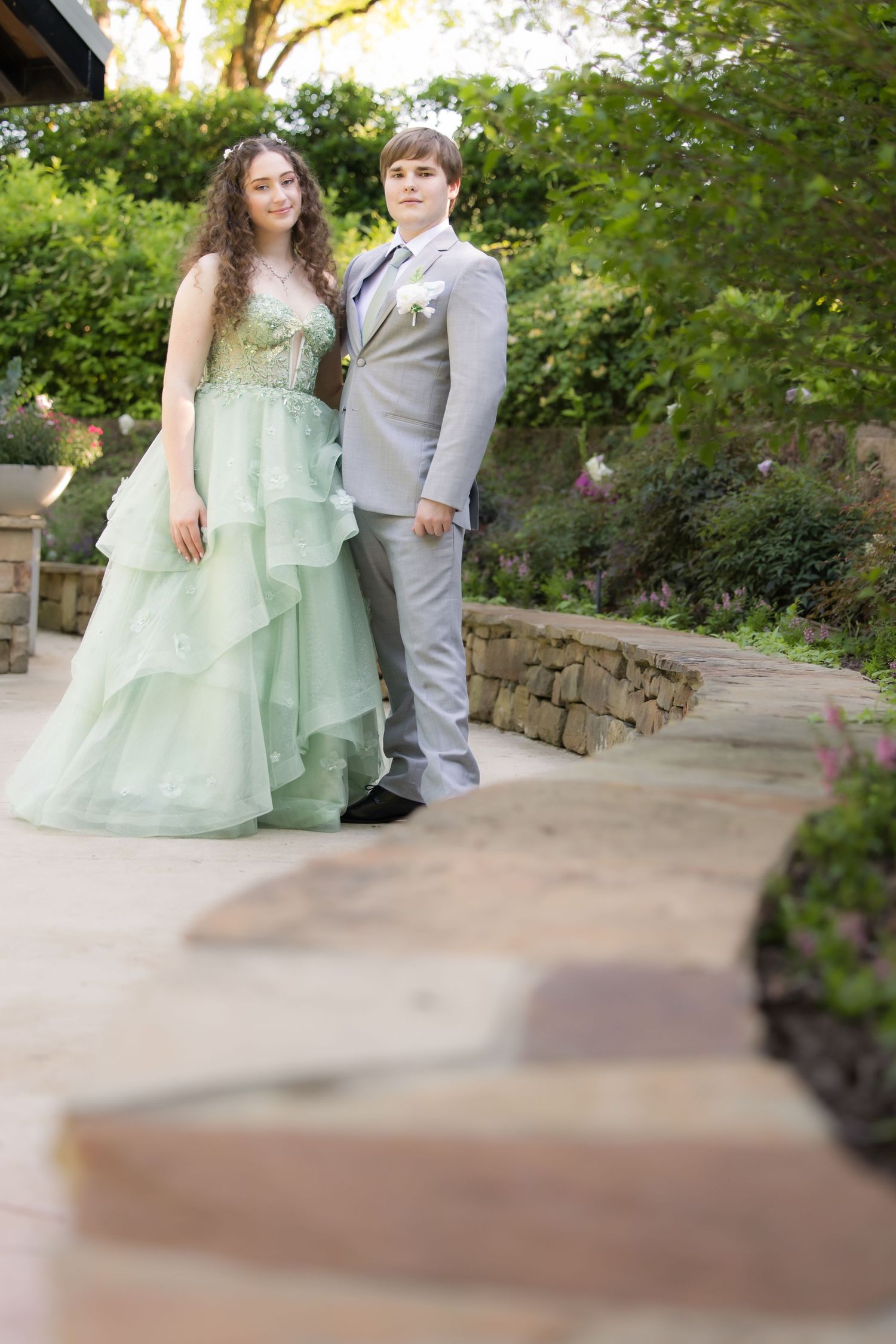 A couple poses on a garden path, with the person in a mint green layered gown and their partner in a light gray suit.