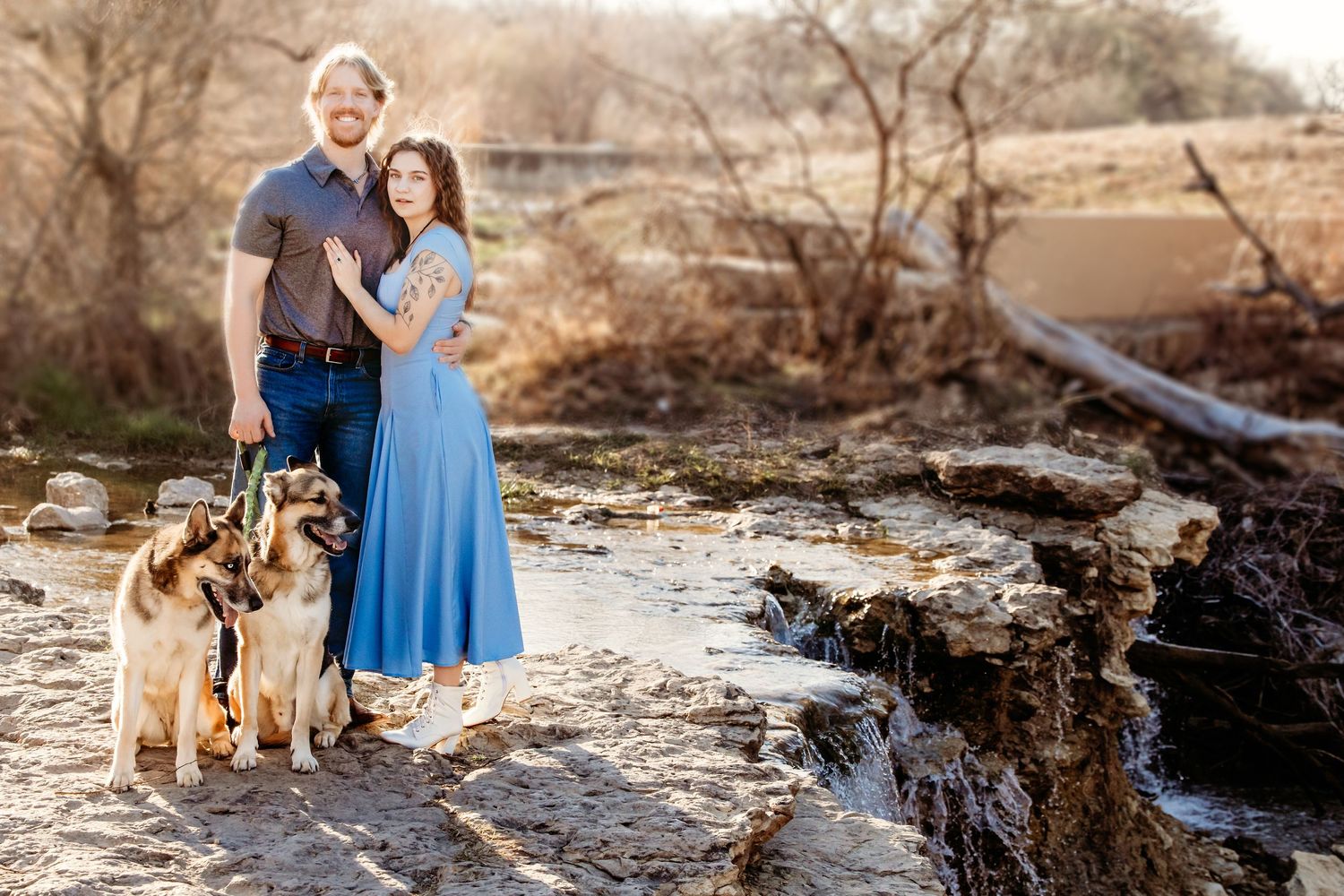 A couple in casual attire stands on rocky terrain near a stream with two golden retriever dogs at sunset.
