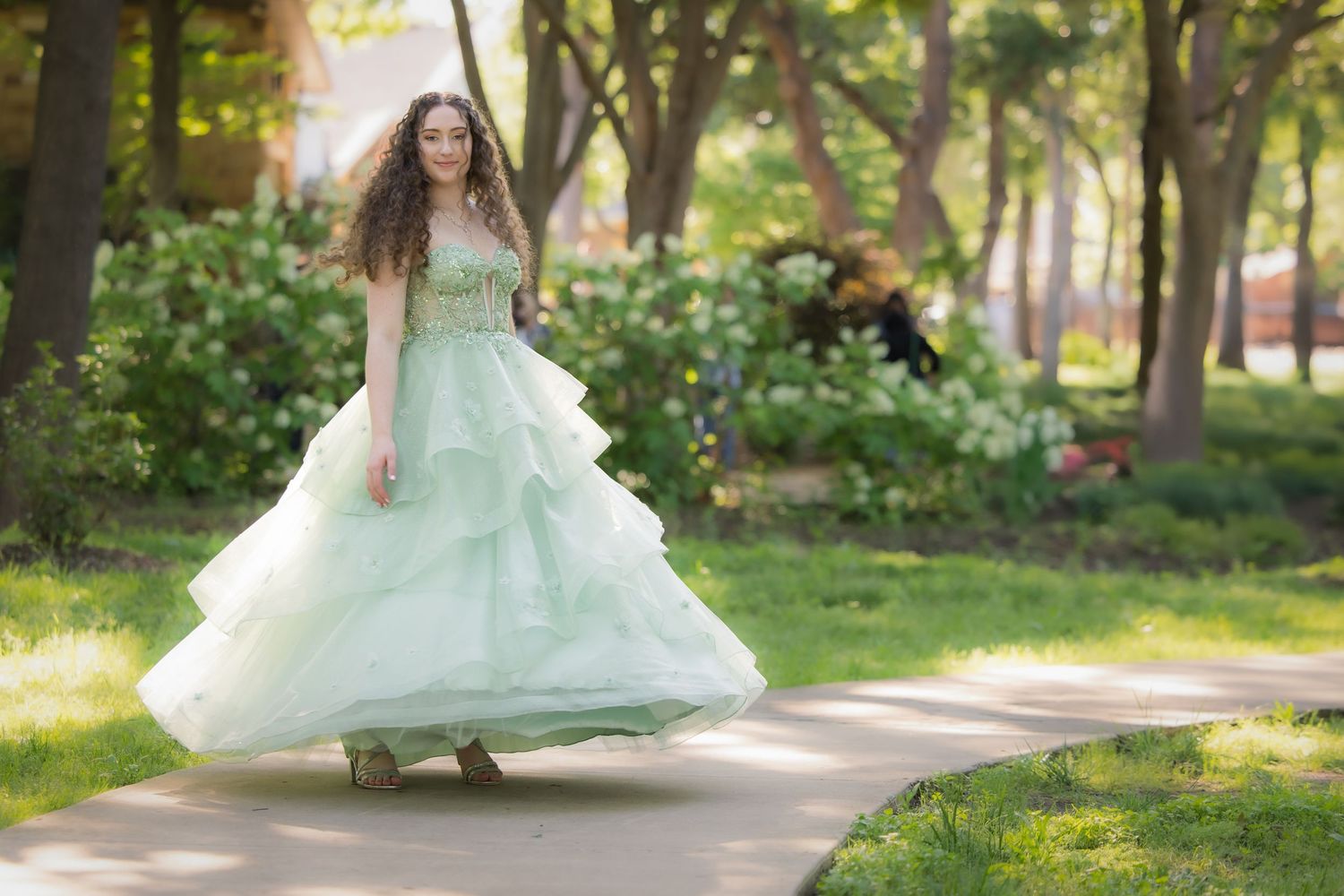 A graceful figure twirls in a mint green tulle gown along a sunlit garden path surrounded by trees.