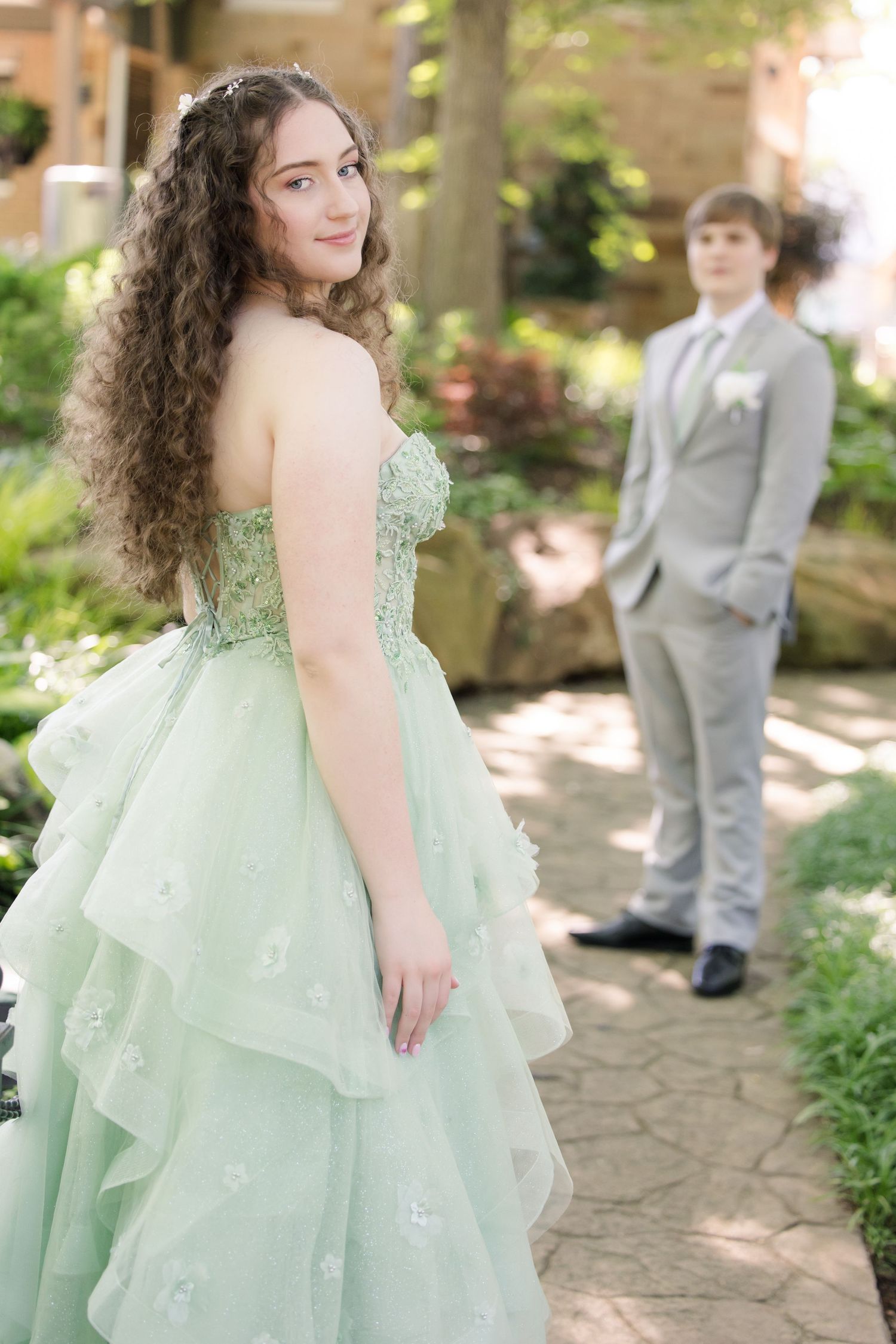 A stunning mint green formal dress stands out in a garden setting while someone in a grey suit waits in the background.