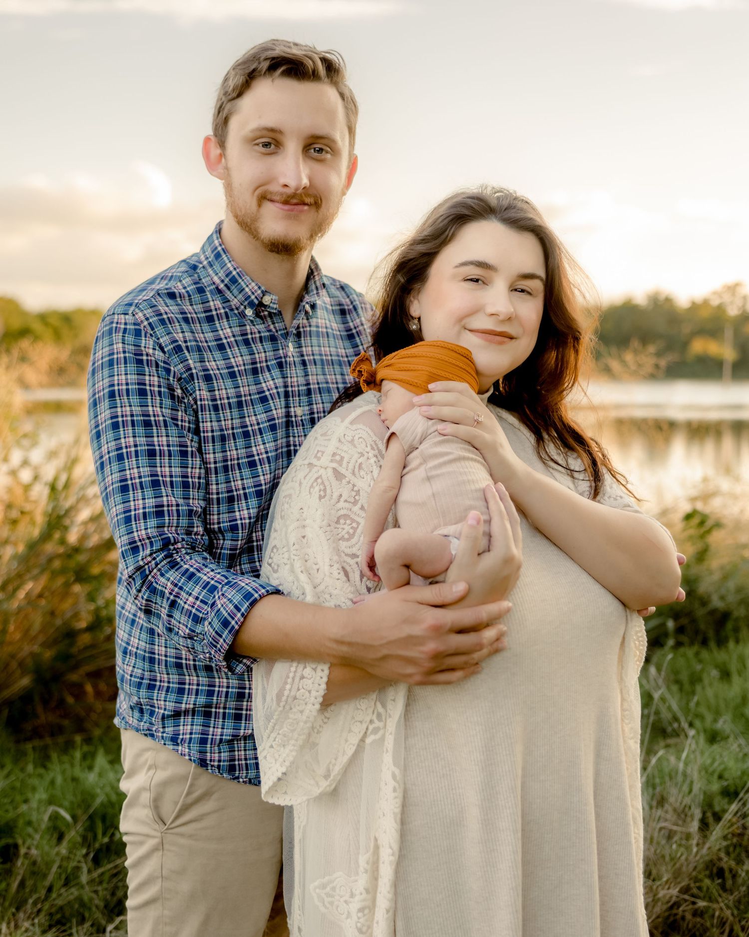 A family of three poses together outdoors at sunset near a lake.