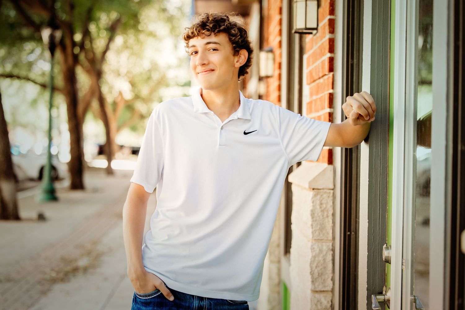 Teen boy in white Nike polo leans against brick building on a sunny urban sidewalk for senior photos.
