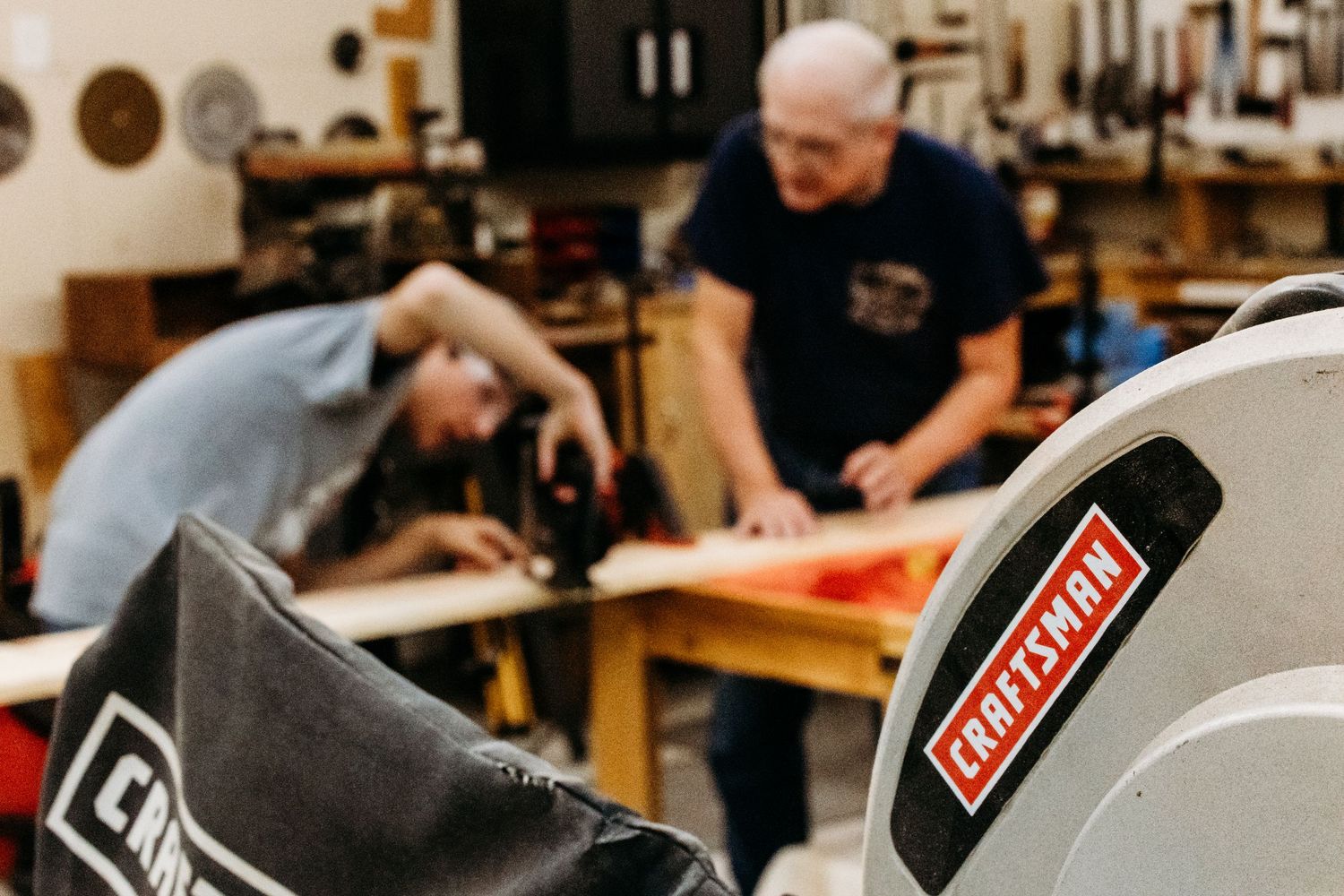Two people working together at a woodworking workbench with a craftsman saw in the foreground.
