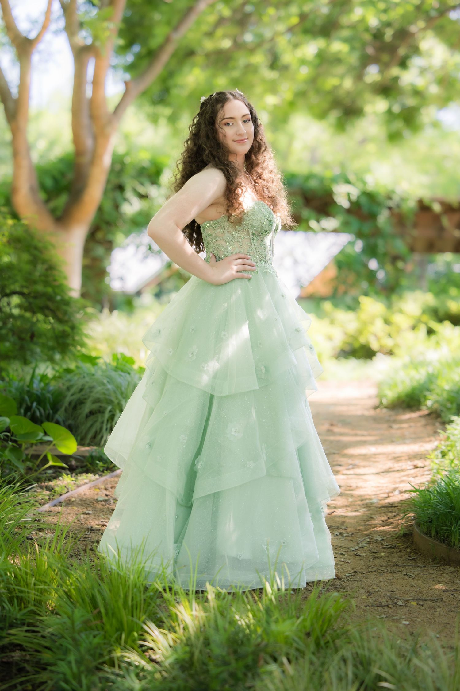 A mint green ball gown with layered tulle skirt photographed on a sunlit garden path surrounded by lush greenery.