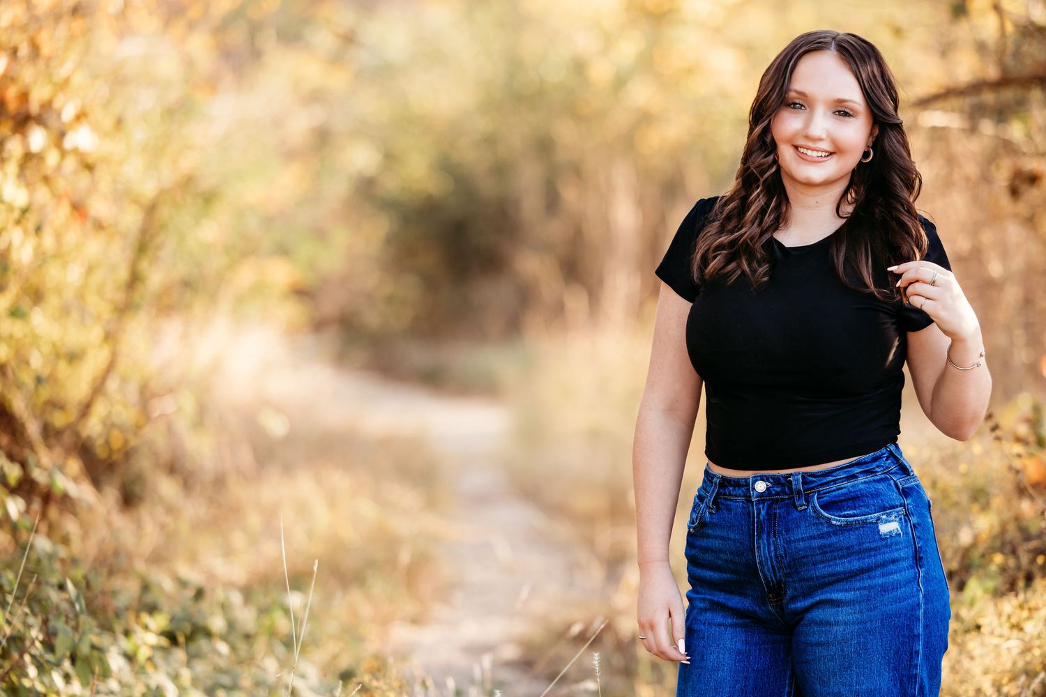 A young lady in a black t-shirt and blue jeans poses on an autumn path with golden foliage in the background in Fort Worth