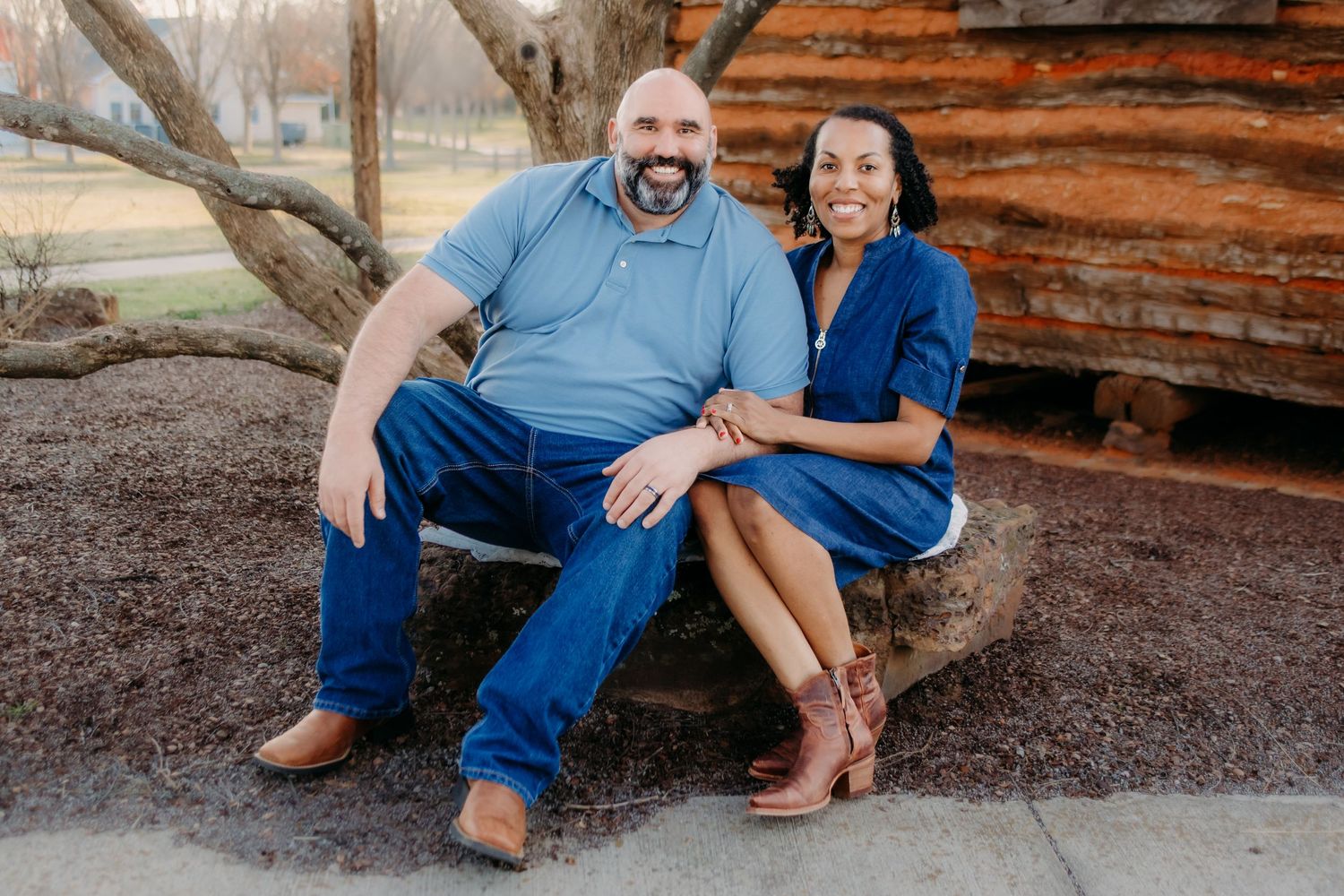 Two people in blue clothing sit together against a rustic wooden cabin in an outdoor setting.