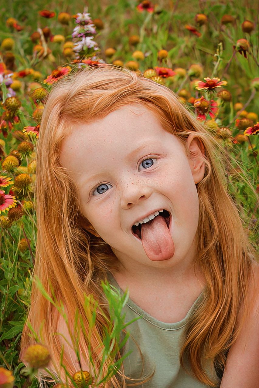 Cheerful redhead girl with blue eyes and freckles sticking out tongue in meadow at Fort Worth, TX family portrait session.