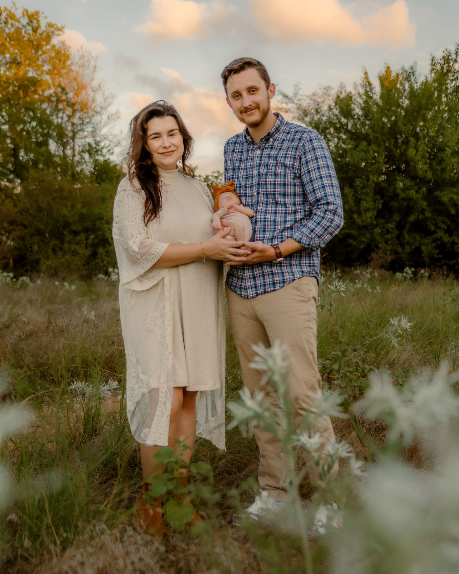 A couple stands together in a field at sunset holding their baby.