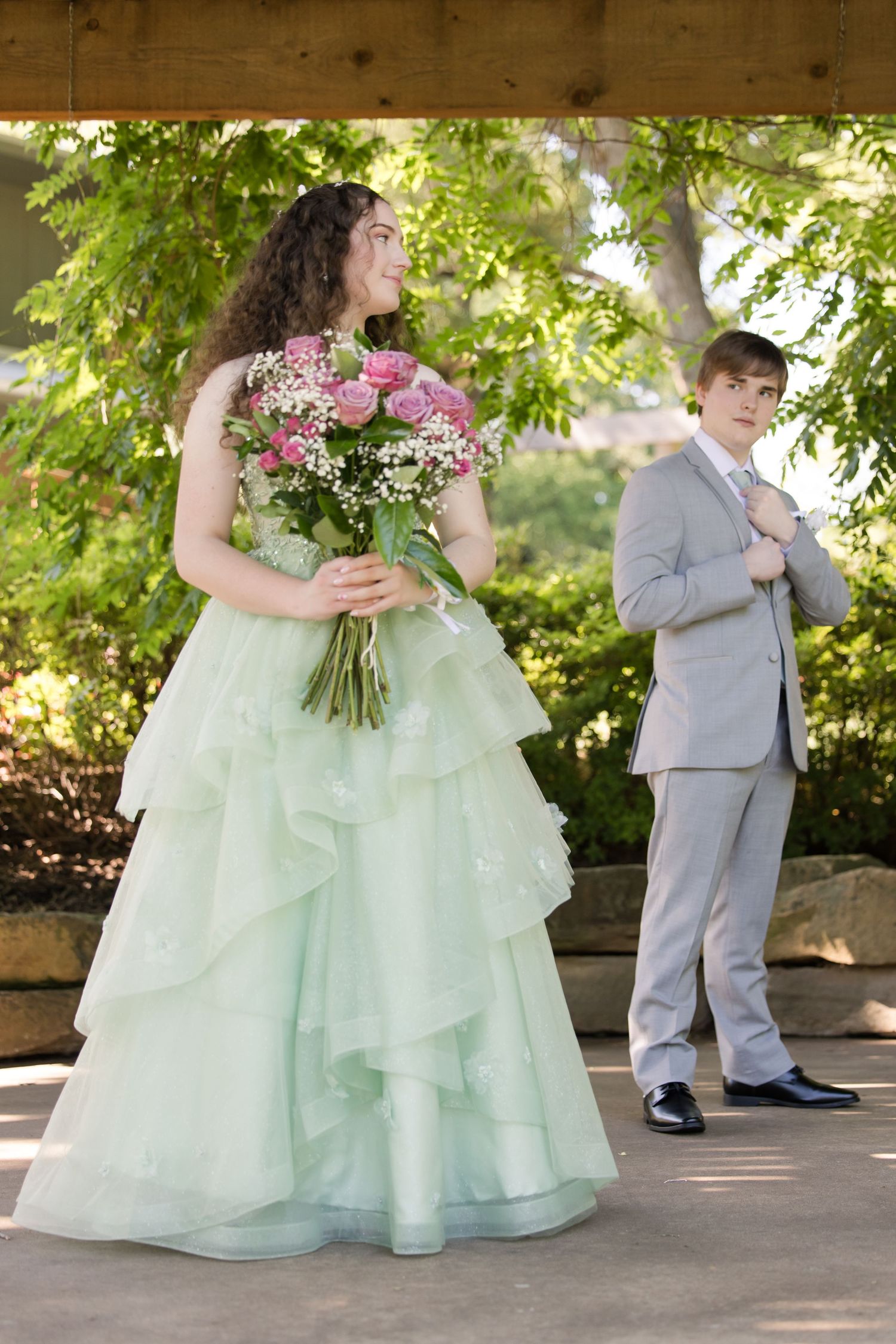 Wedding couple in mint green gown and gray suit pose outdoors under trees with pink flower bouquet.