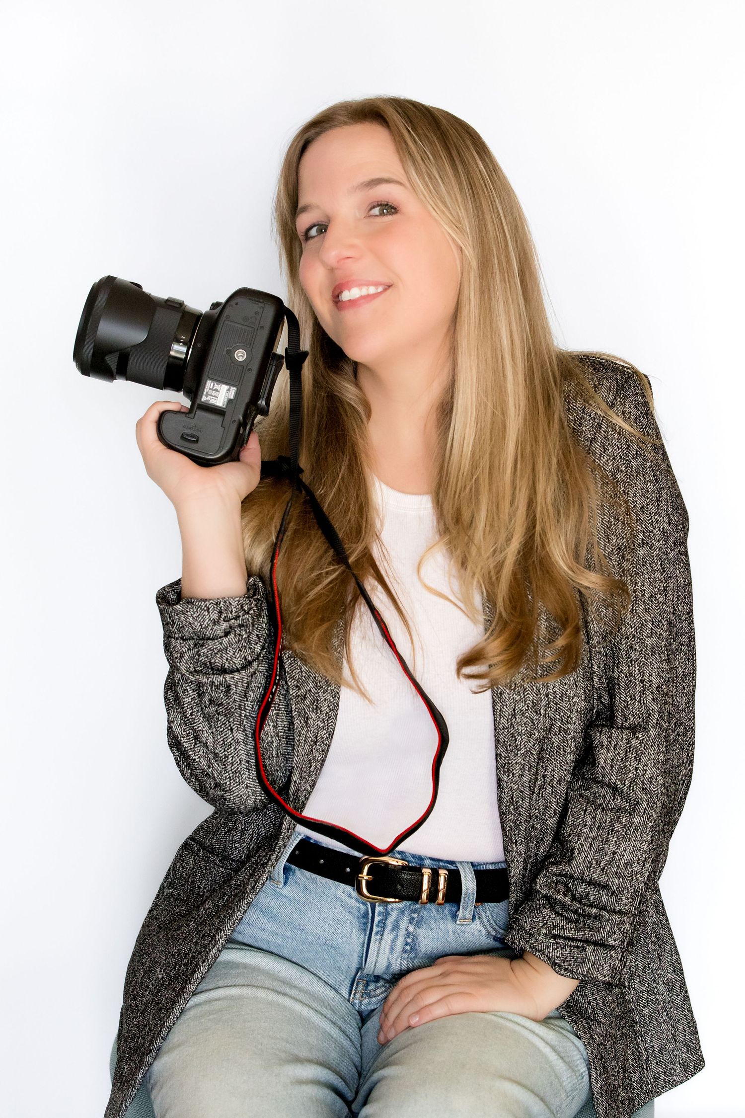 Blonde female photographer holding a DSLR camera, wearing a gray blazer and jeans, smiling against a white background.