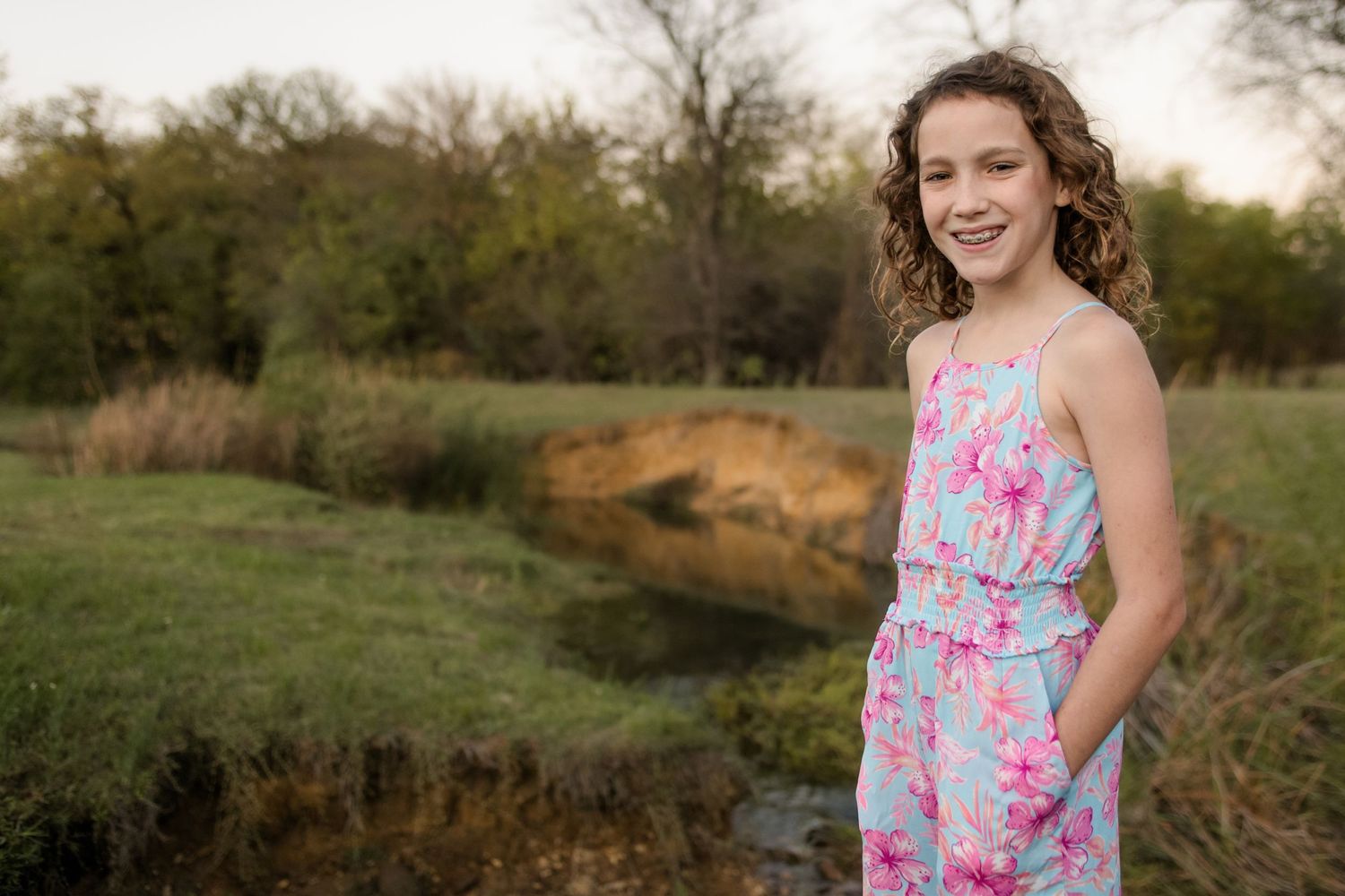 Young person in a floral summer dress poses outdoors by a pond at sunset with trees in the background.