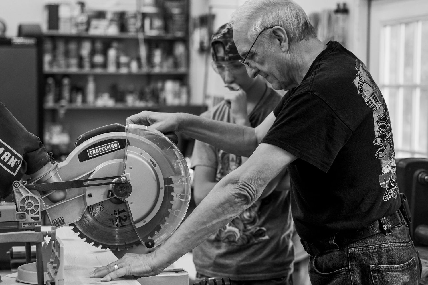 Black and white photograph of woodworkers operating a miter saw in a workshop with tools and supplies visible.