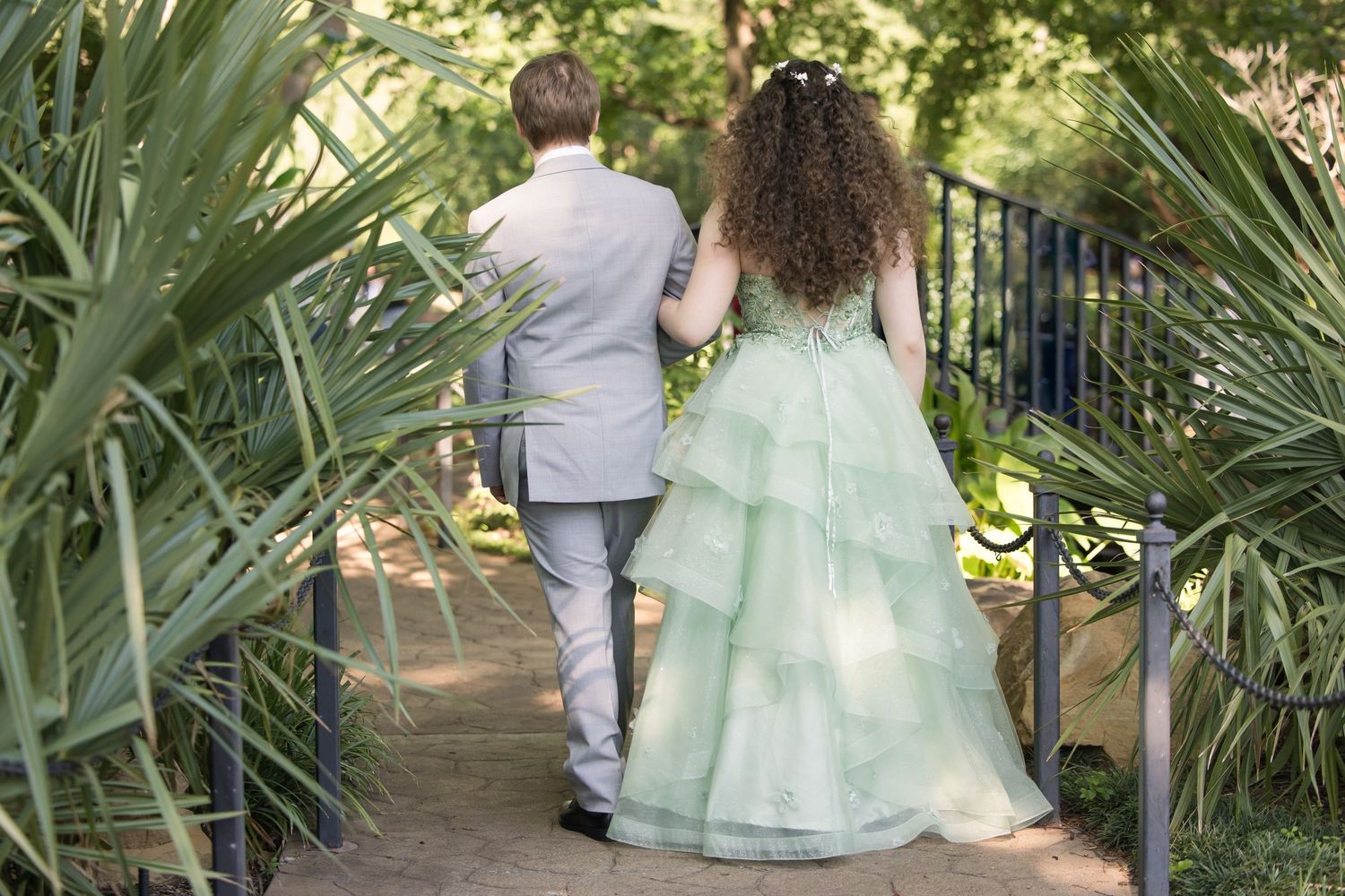 A couple walking together on a garden path surrounded by tropical palm fronds, with the bride wearing a mint green gown.