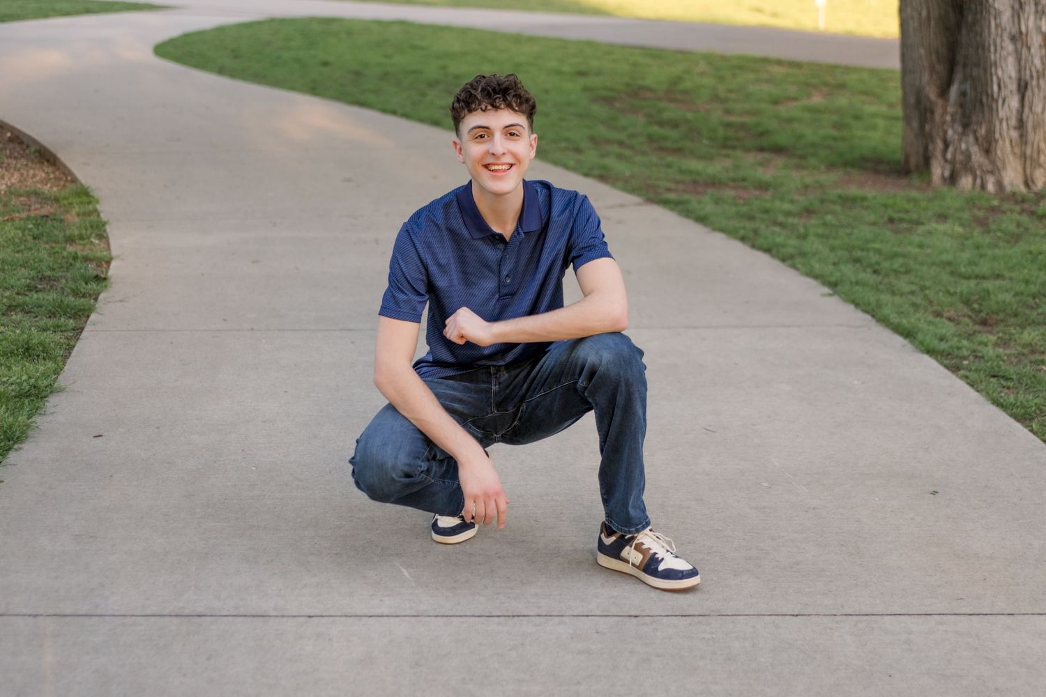 Person in navy blue shirt and jeans crouches on a curved sidewalk path surrounded by green grass.