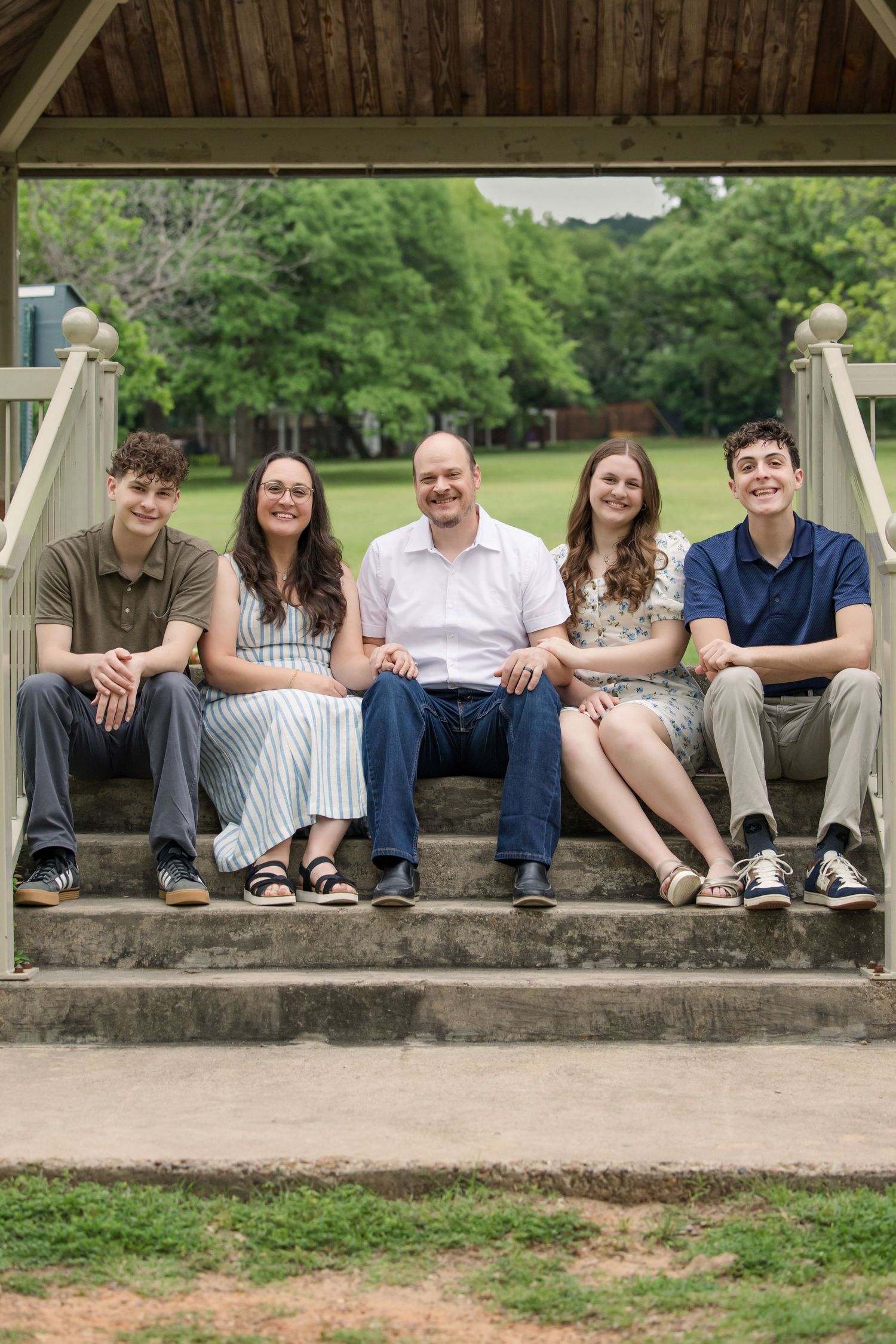Five people sit together on outdoor stone steps with trees and greenery visible in the background on a sunny day.