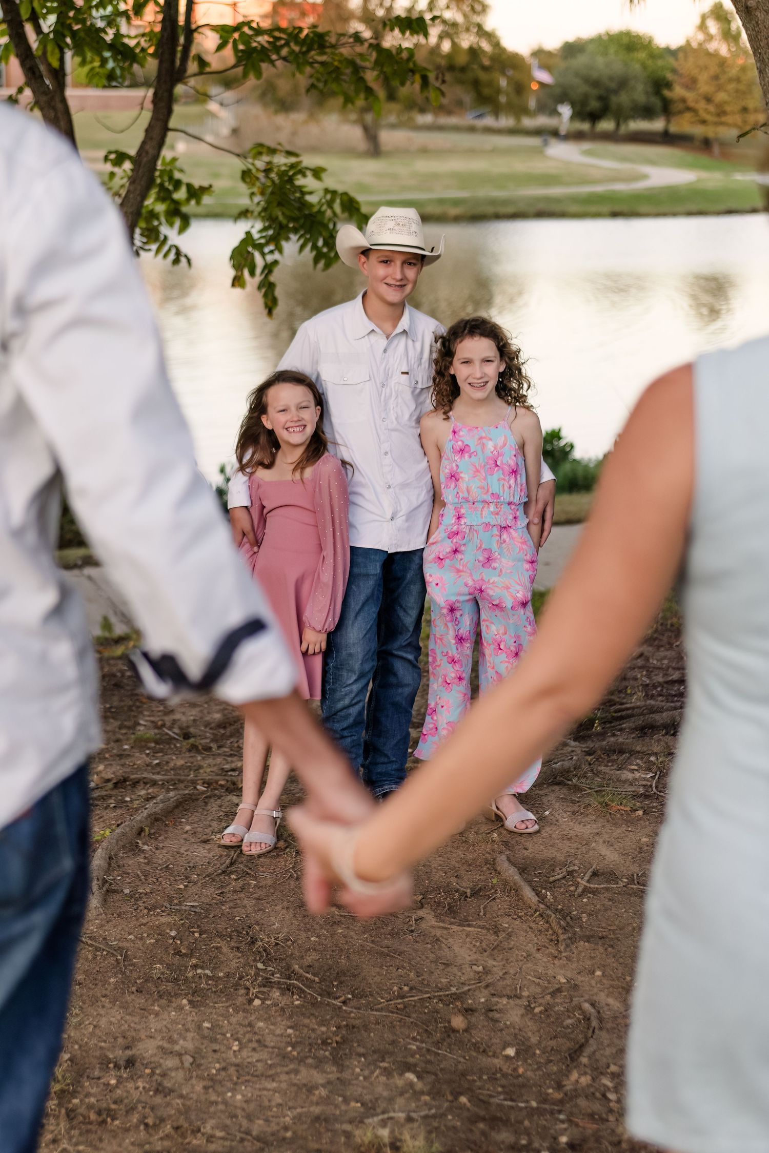 A family poses near a lake at sunset with blurred foreground hands framing the composition.