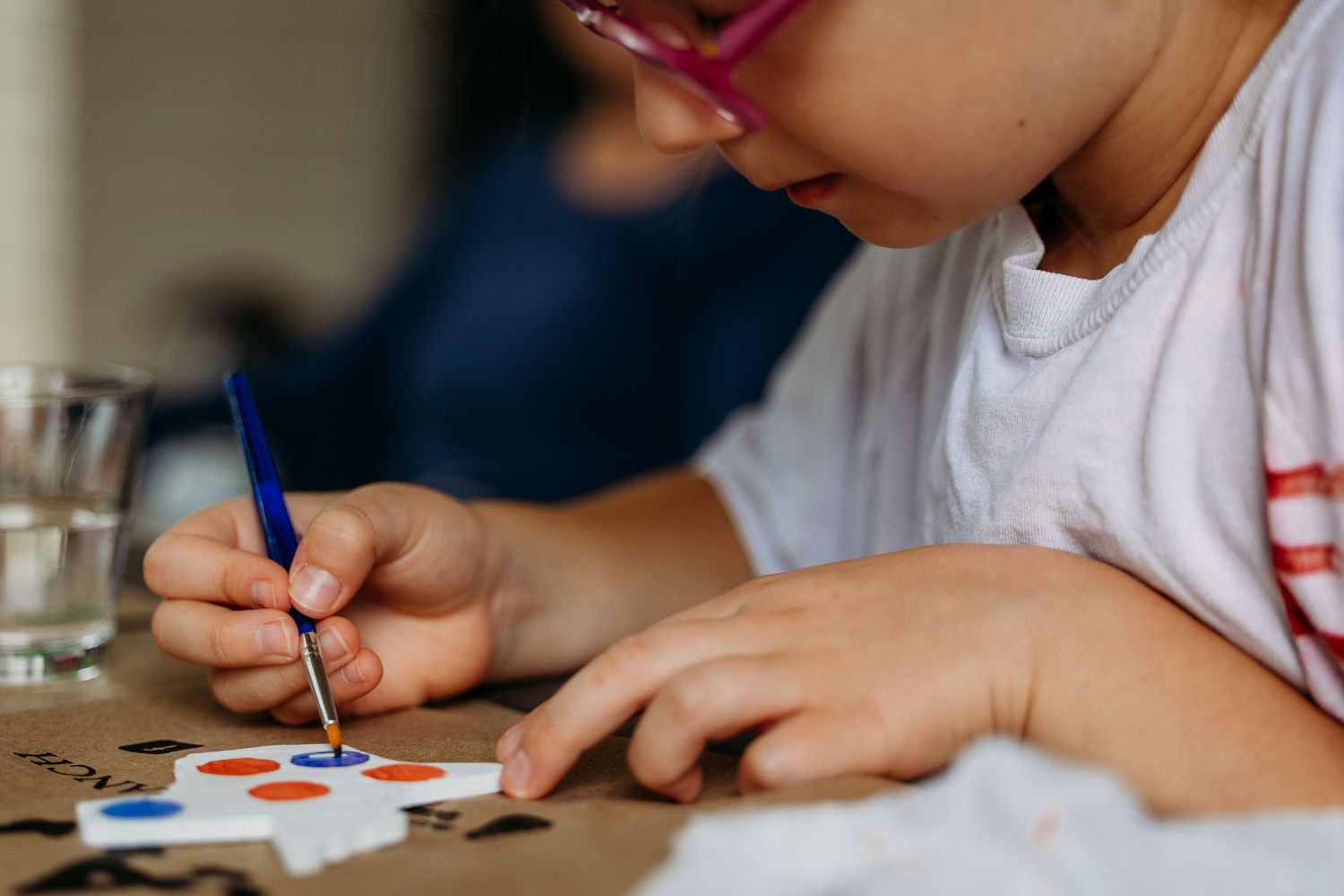 Someone wearing pink glasses uses a blue pen to carefully paint or draw details on a small surface.