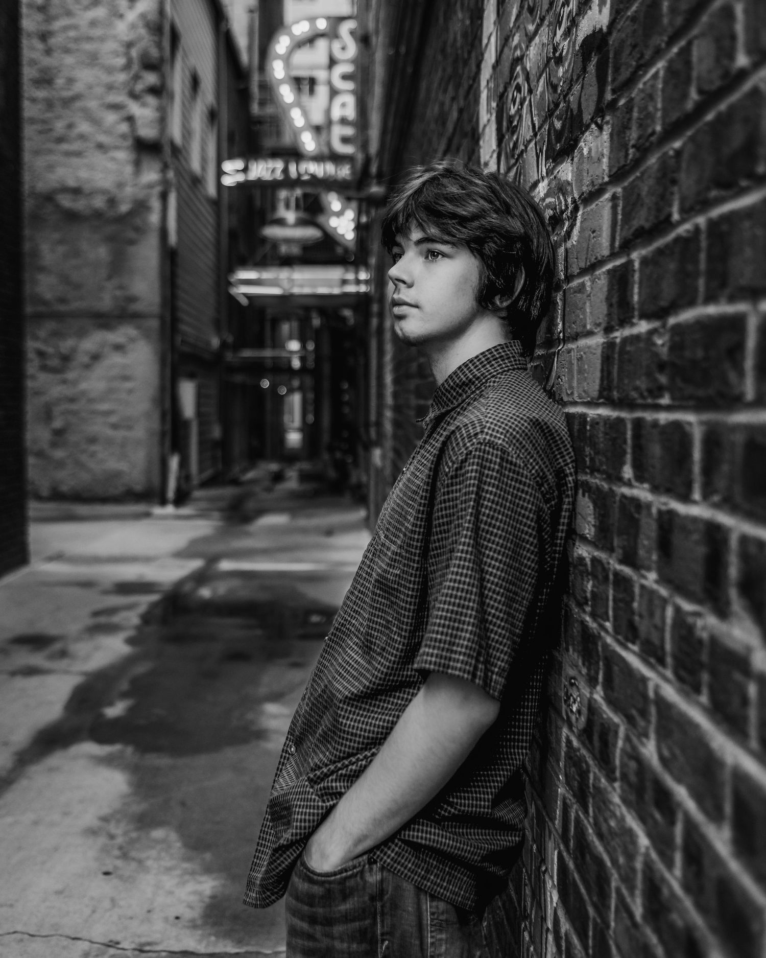 Black and white portrait contemplative figure leaning against brick wall in urban alleyway setting.