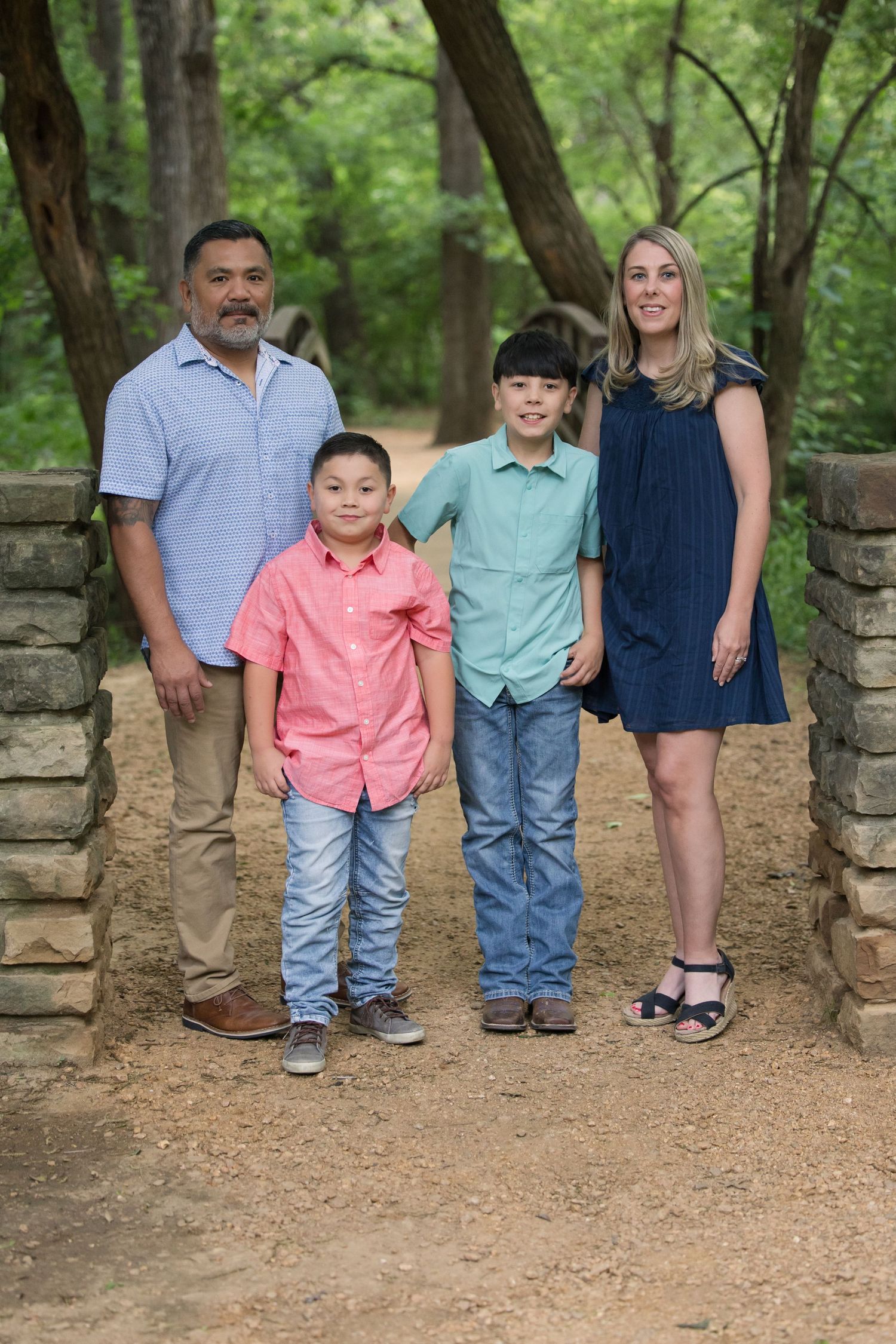 A family stands together on a dirt path surrounded by green trees and a stone wall in a natural outdoor setting.