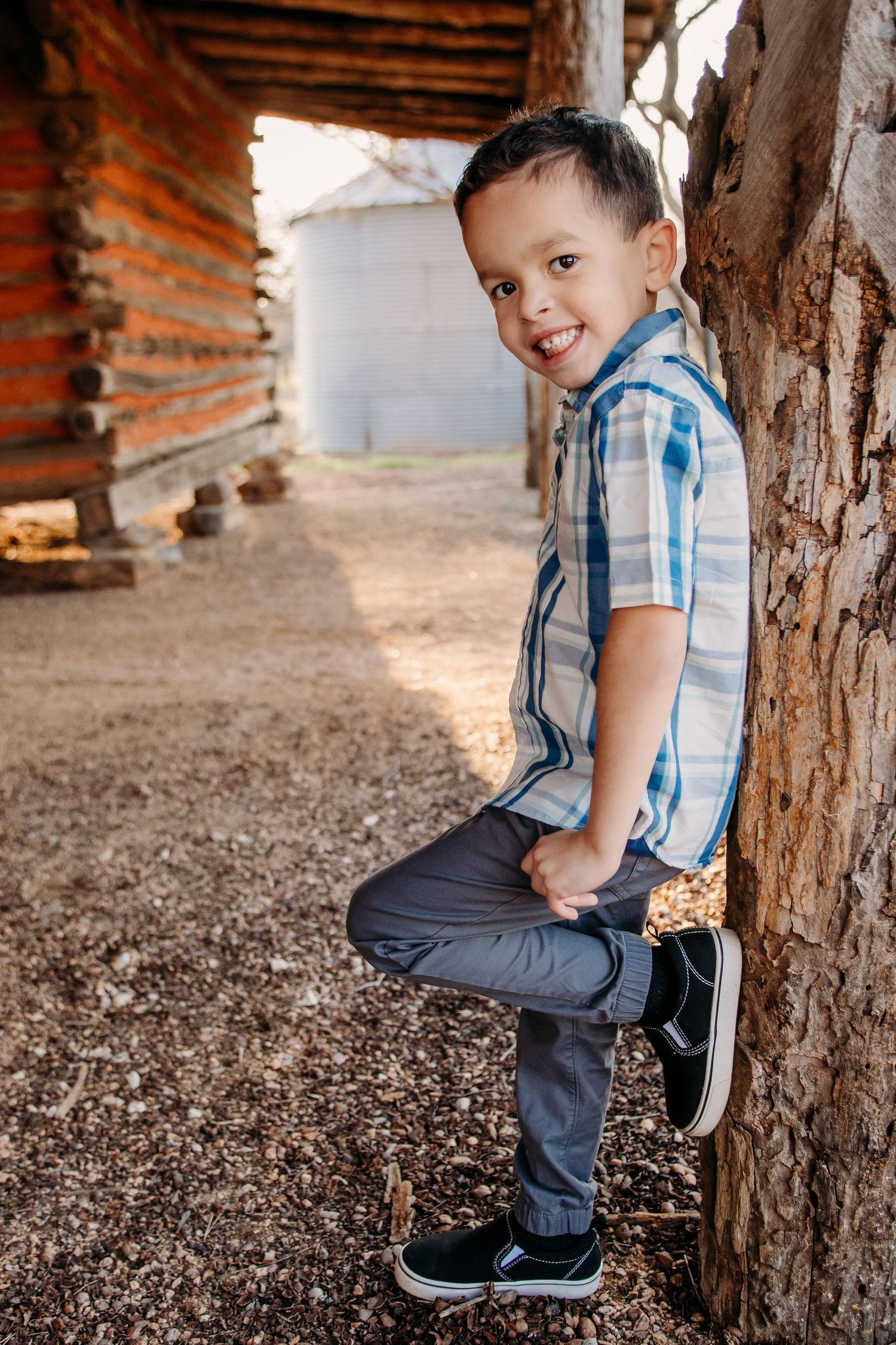Child in blue plaid shirt and jeans leans casually against rustic brick wall in outdoor setting.