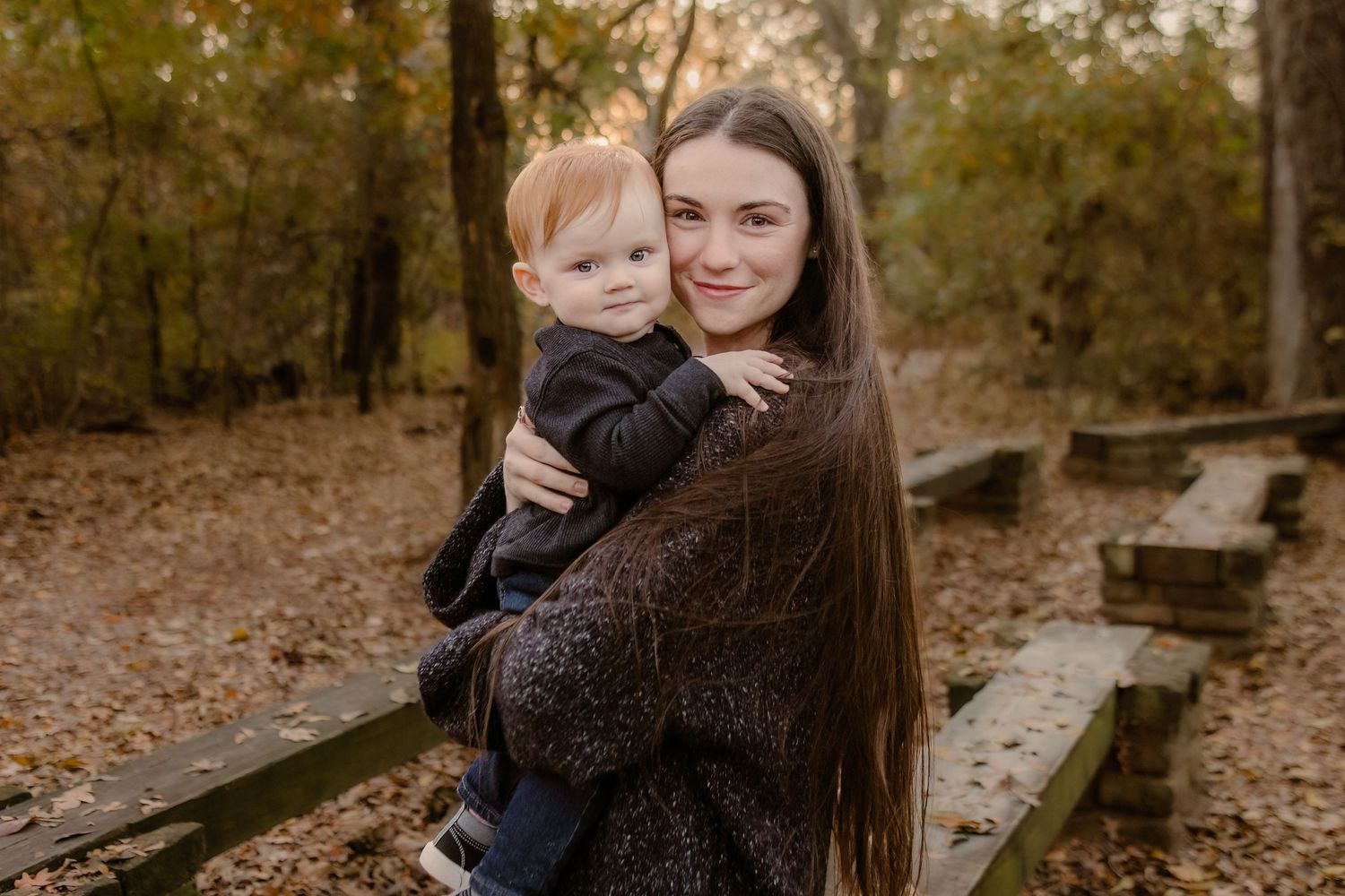 A woman with long dark hair holds a baby while standing on wooden benches in an autumn forest setting.