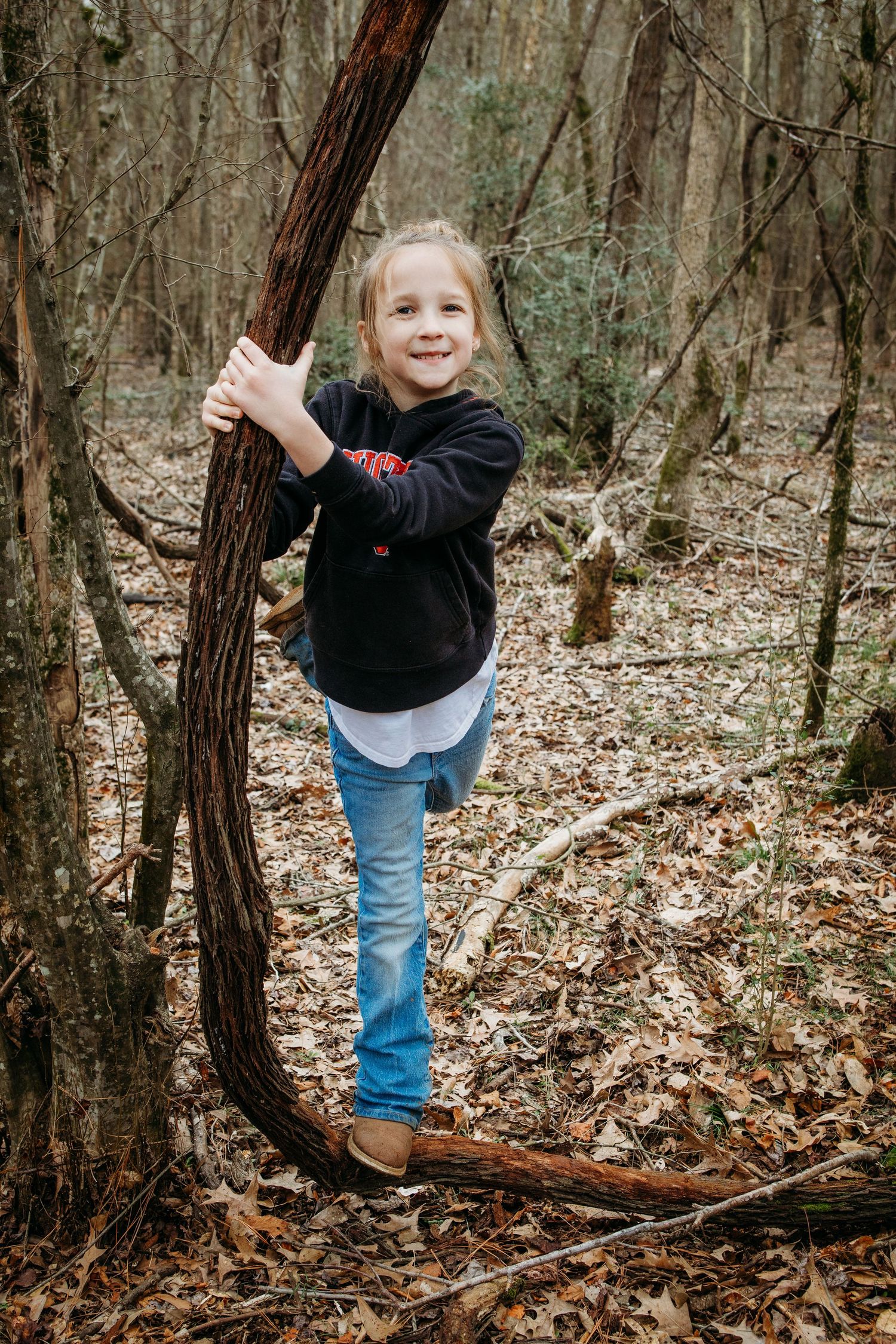 A child plays and explores in a forest setting filled with fallen leaves and twisted tree trunks.