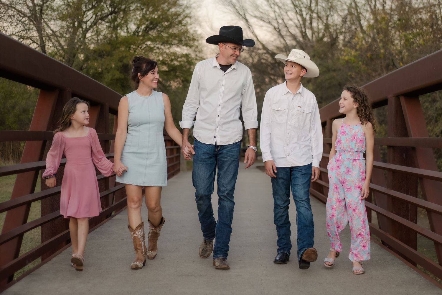 Family of five walks together in casual attire on a wooden bridge during sunset.
