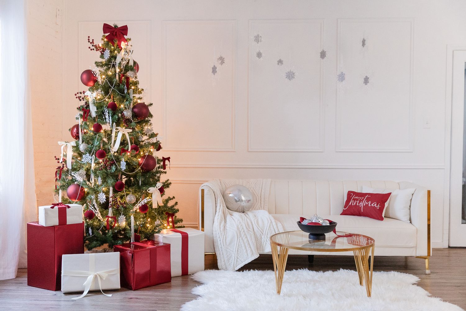A decorated Christmas tree with red presents stands beside a white sofa and gold coffee table in a modern living room.