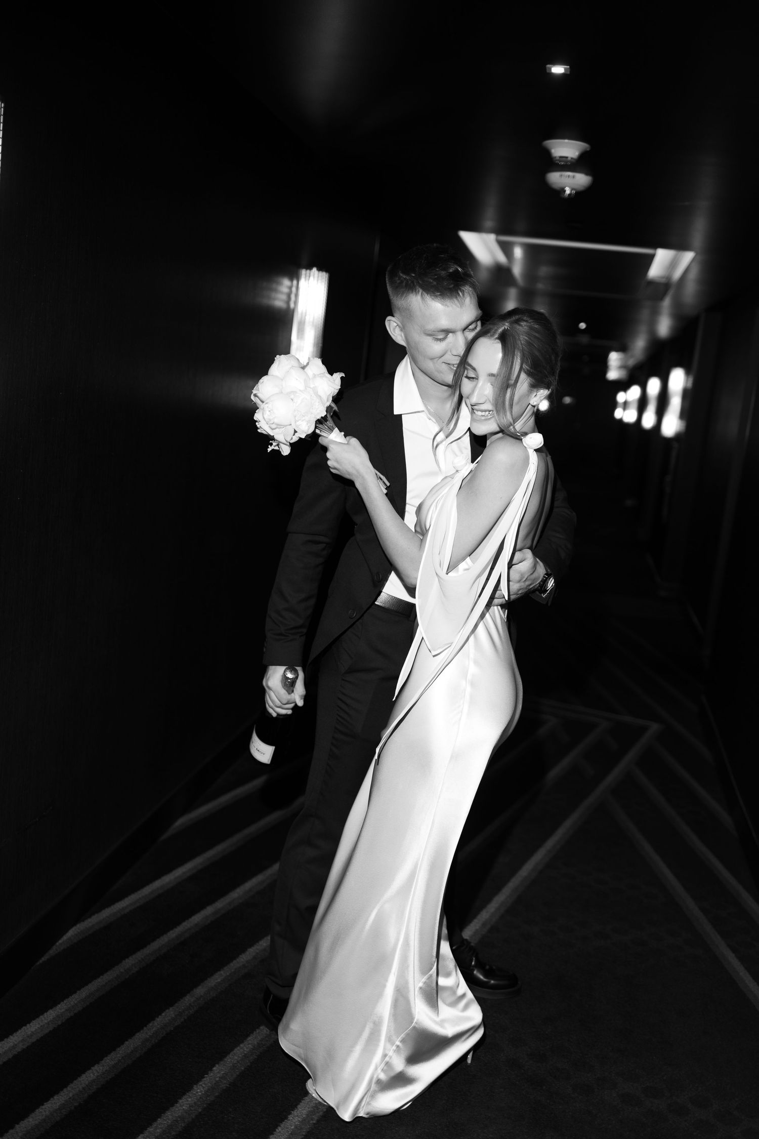 A couple in wedding attire share an intimate dance moment in a dark hallway with a white bouquet.