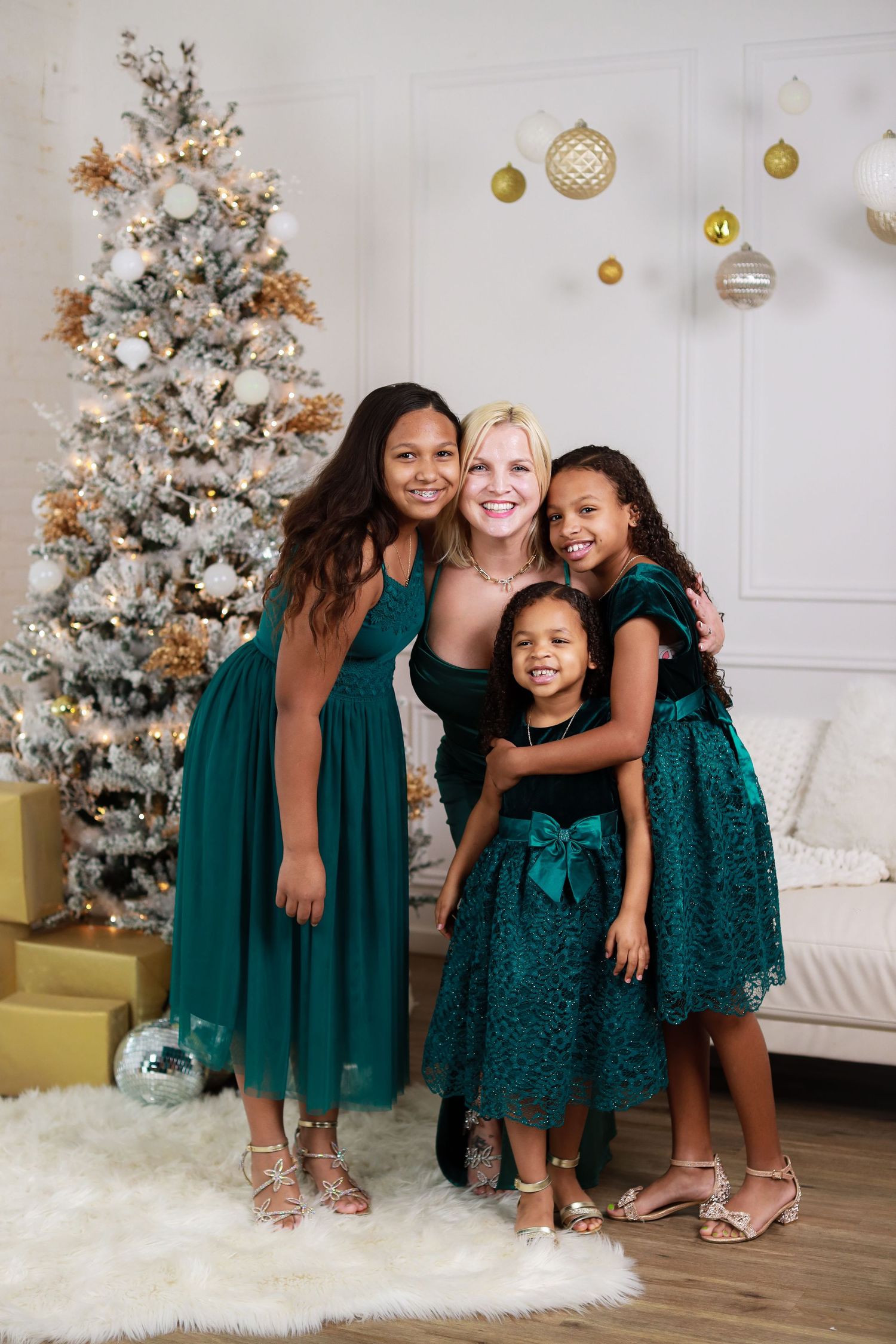 Young friends in emerald green dresses share laughter and hugs by a silver Christmas tree during holiday photos.