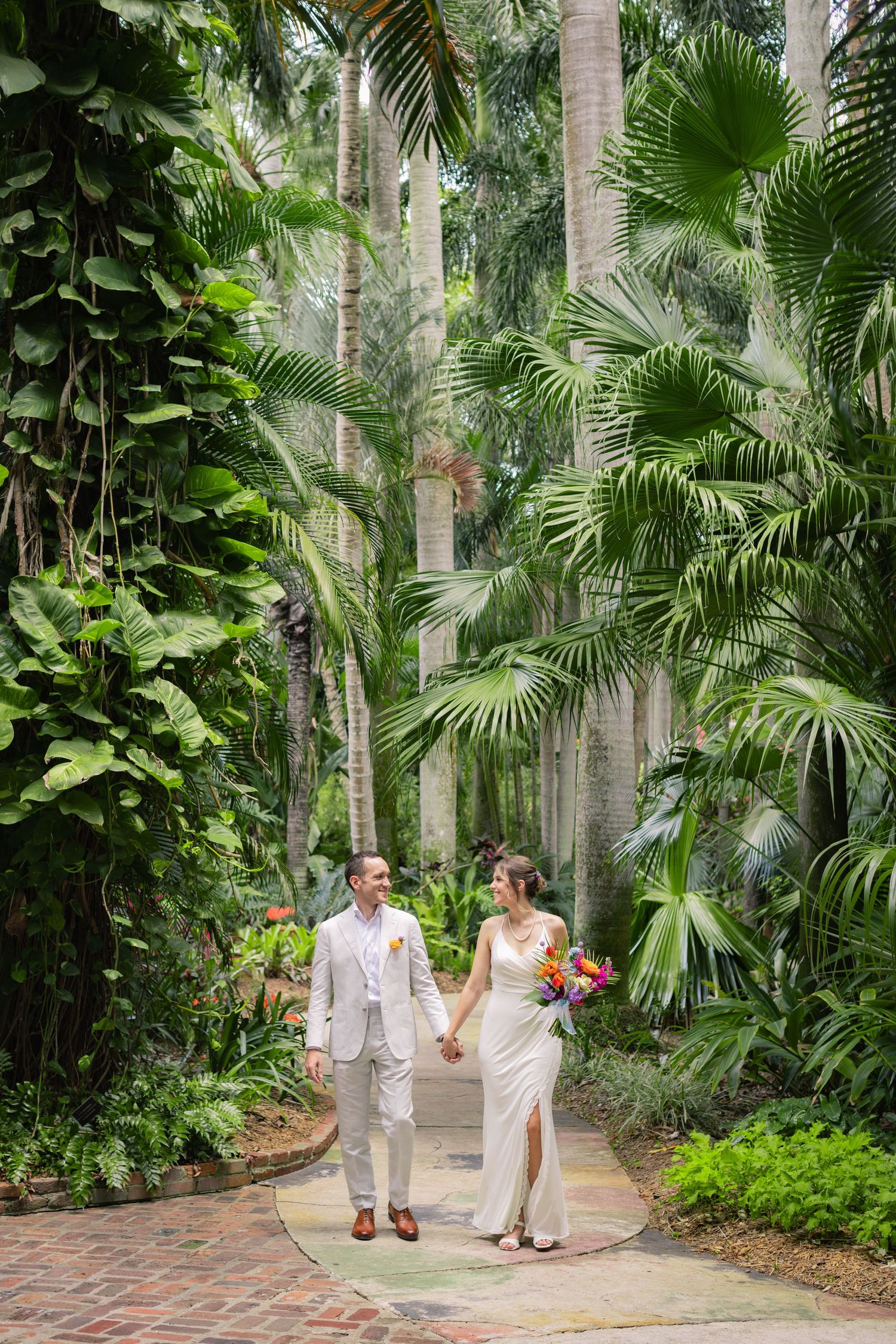 Newlyweds walk hand in hand along a tropical garden path surrounded by lush palm trees and foliage.