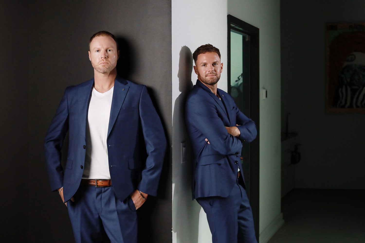 Two men in blue business suits pose confidently against a modern interior wall.