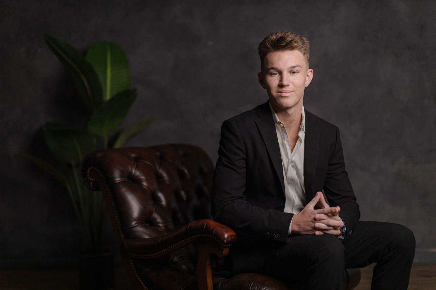Professional portrait of a young man in dark blazer seated by vintage leather chair.