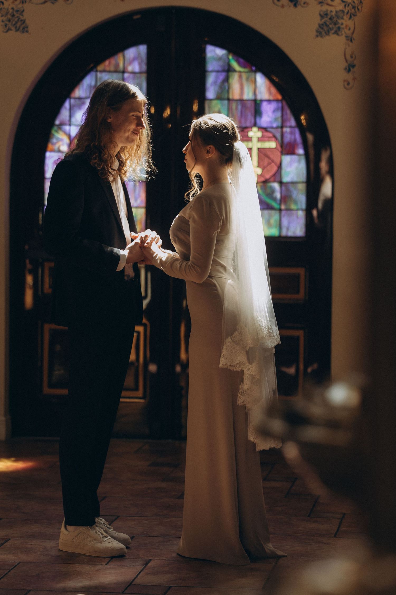 A couple exchanges wedding vows in front of a stained glass window in a dimly lit church setting.