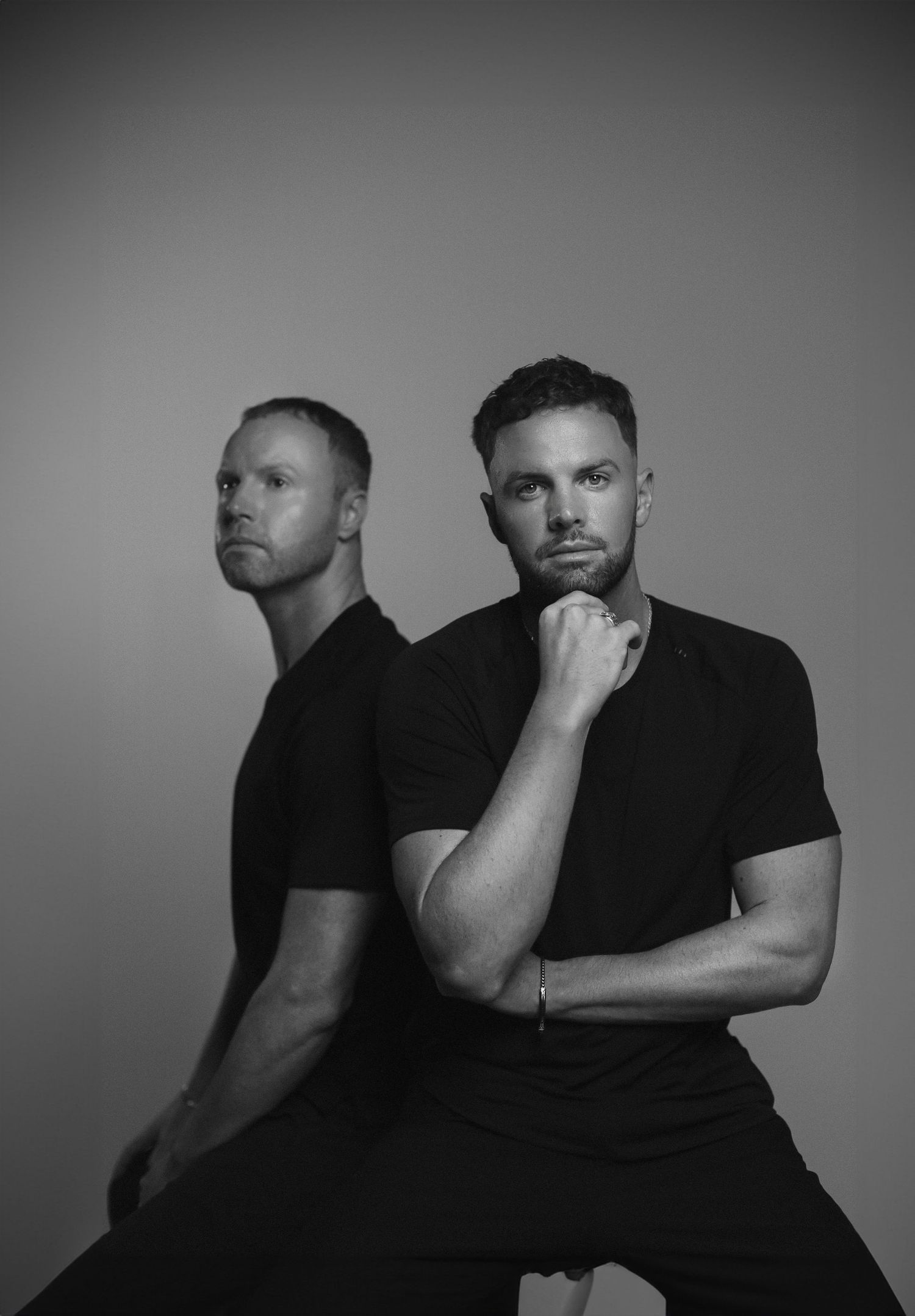 Artistic black and white portrait of two men in dark clothing with dramatic lighting.