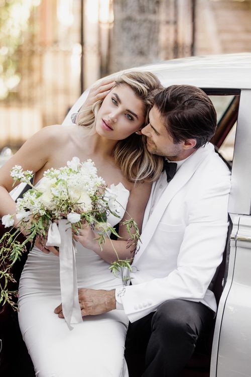 Romantic wedding couple embracing by vintage car with elegant floral bouquet.