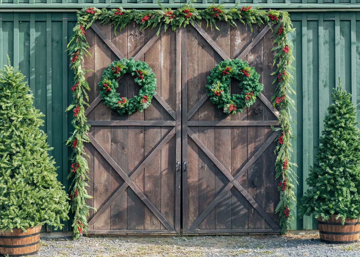 Rustic wooden barn doors with green garland, red berries and two Christmas wreaths, flanked by potted evergreen trees on a green wall.