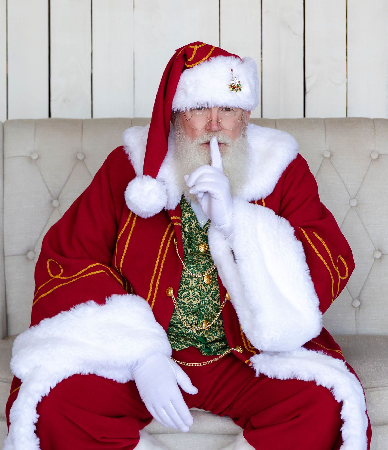 Santa Claus in red suit sitting on white chair making a shush gesture with white gloved finger to lips.
