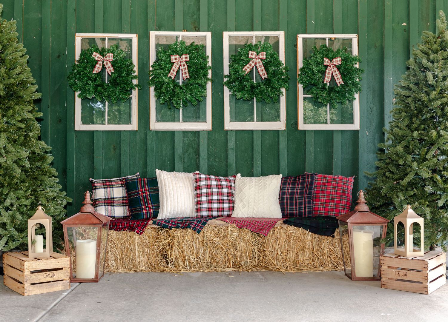 Rustic Christmas photo backdrop with a hay bale bench, plaid pillows, evergreen wreaths on a green barn wall, trees, and lanterns.