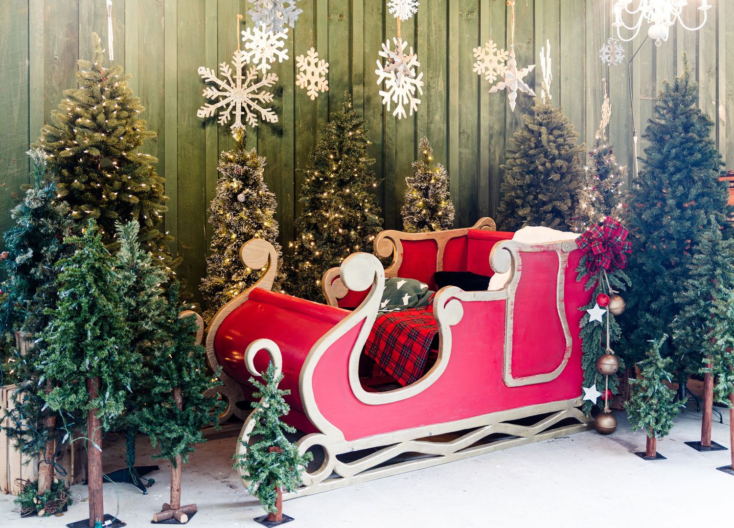 Red and white Santa sleigh with plaid cushions surrounded by small Christmas trees and snowflake decorations against a green wooden wall.