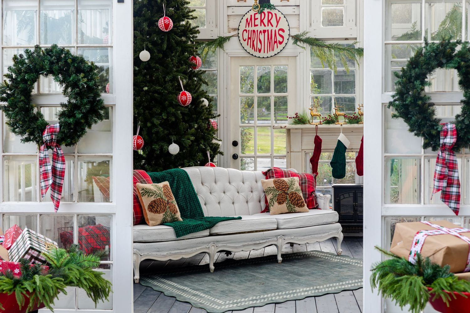 Cozy sunroom decorated for Christmas with a tufted white sofa, plaid pillows, evergreen wreaths, a tree and a Merry Christmas sign.