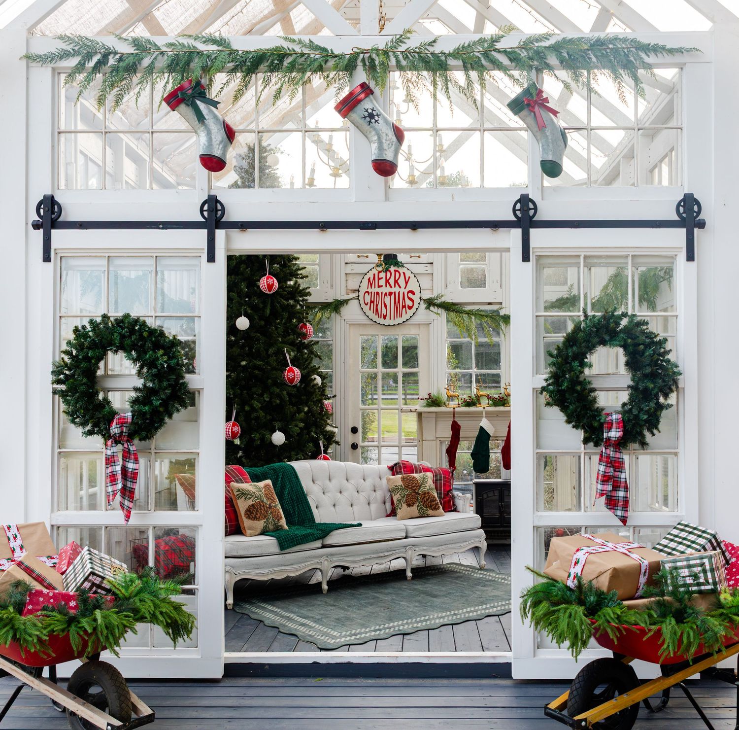 Festive Christmas sunroom with barn doors, evergreen wreaths, hanging stockings, a decorated tree, and a cozy tufted sofa.