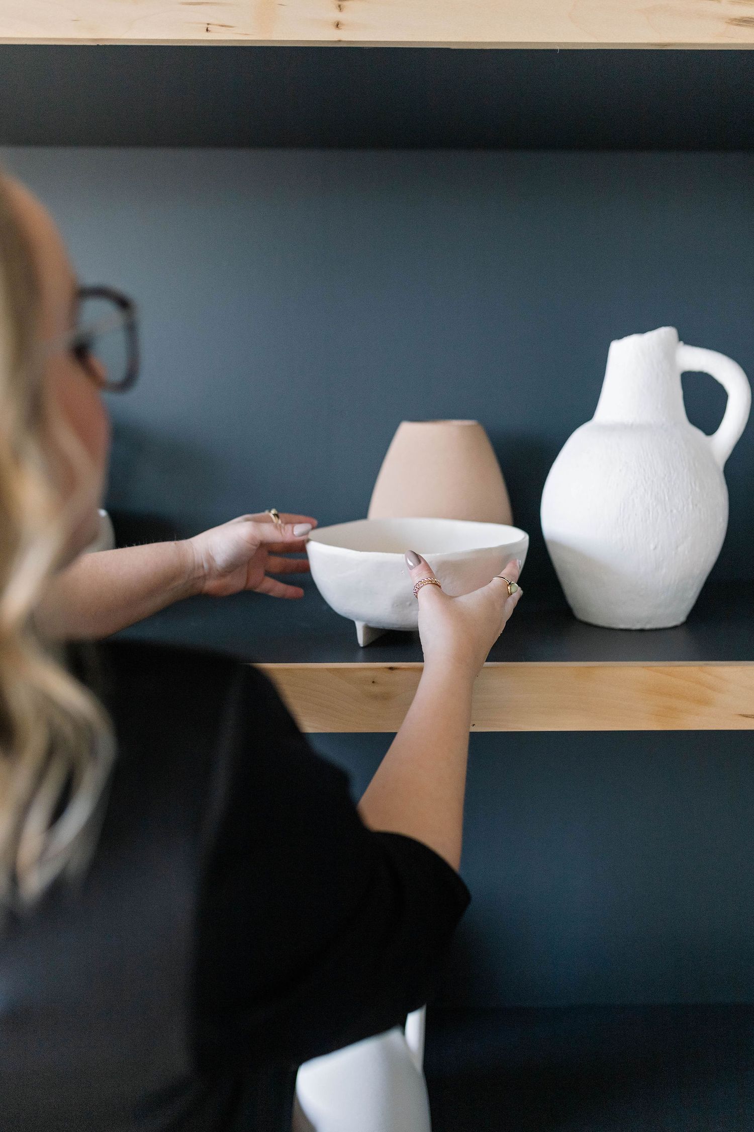 Someone reaching for a white ceramic bowl on wooden shelf displaying minimalist pottery pieces in muted tones.